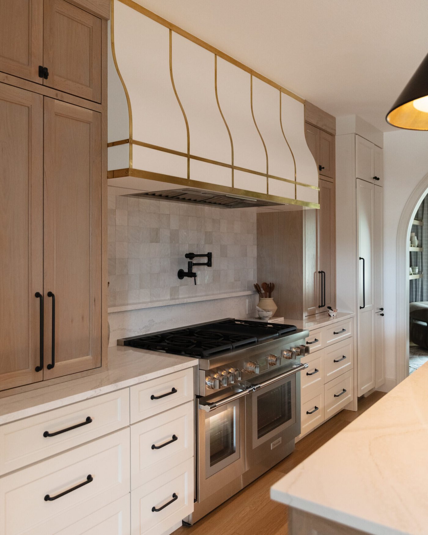 Range wall with a brass-strapped hood, white oak cabinetry and a stone slab backsplash in the Alexandria project by Jennifer Kizzee Design