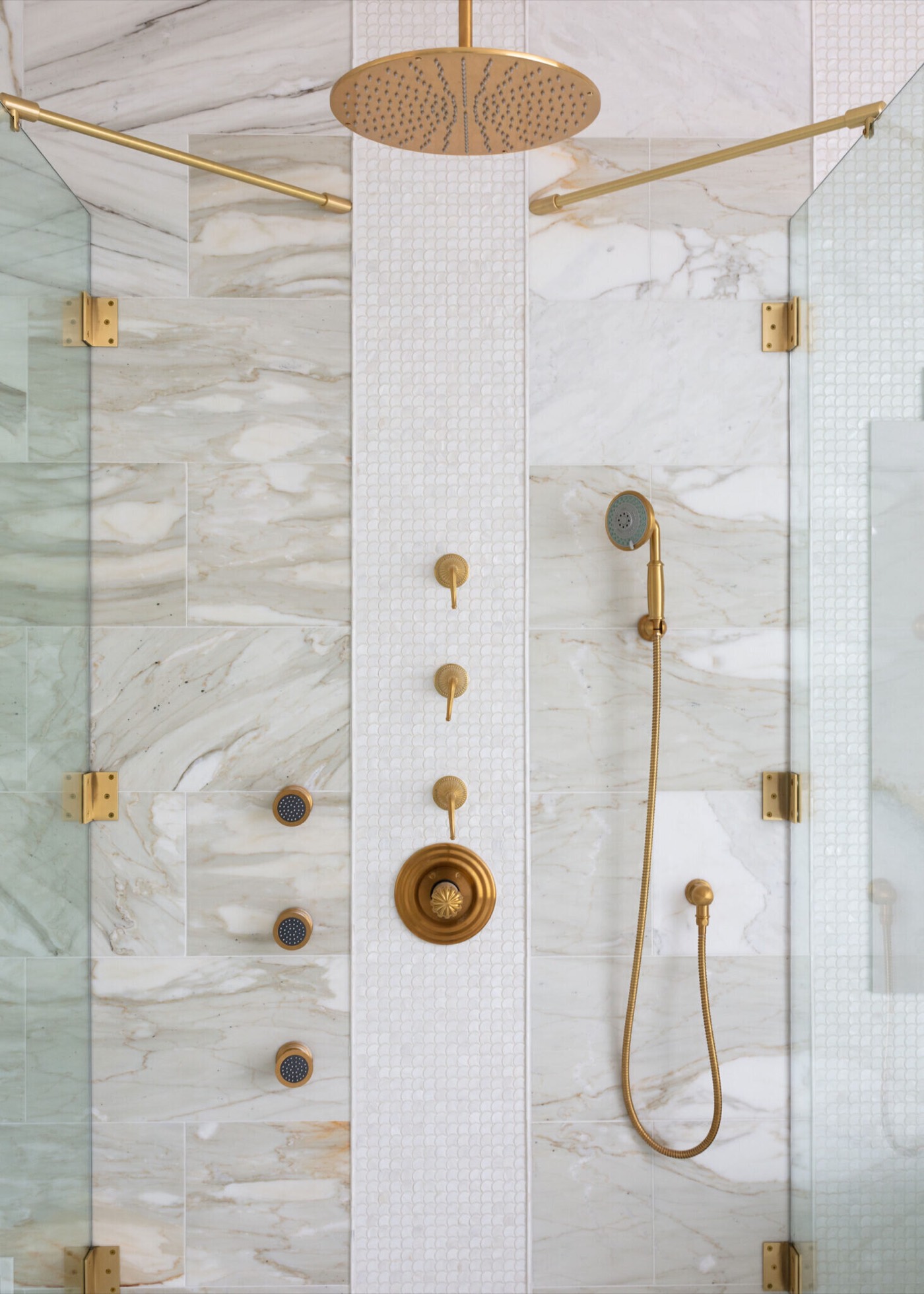 The shower interior with a brass rainhead, ornate thermostatic valve trim and handshower set against white and gold marble split by a floor-to-ceiling mosaic stripe in the Blue Heron Bathroom project by Jennifer Kizzee Design