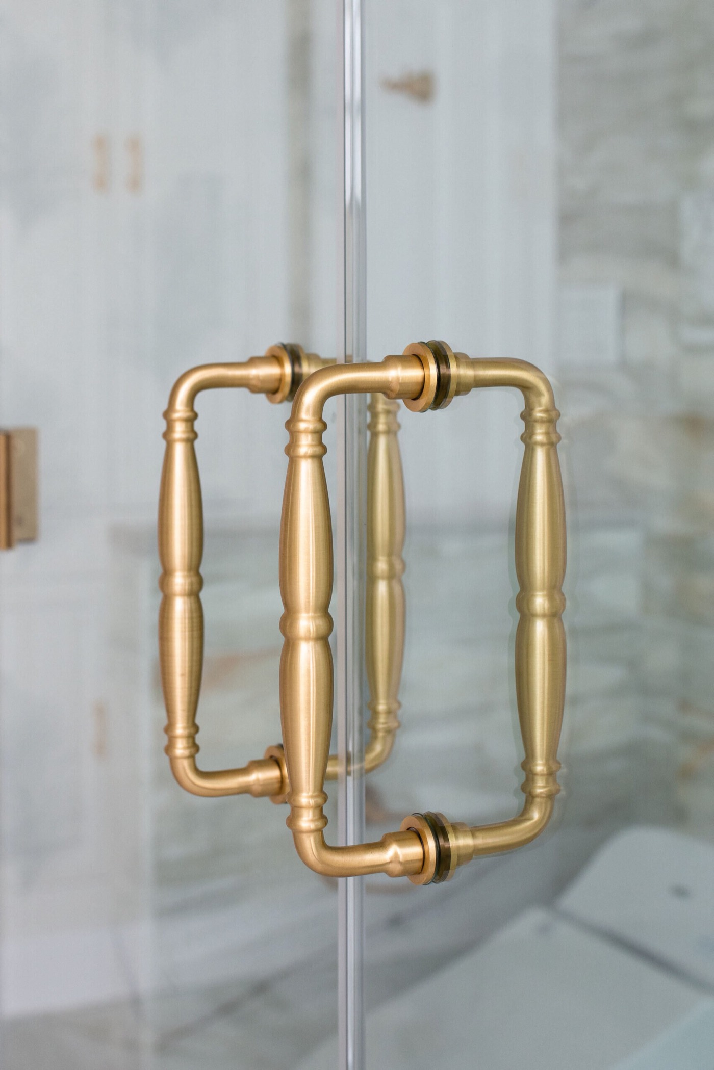 Back-to-back turned satin brass shower door pulls on clear frameless glass with marble beyond in the Blue Heron Bathroom project by Jennifer Kizzee Design