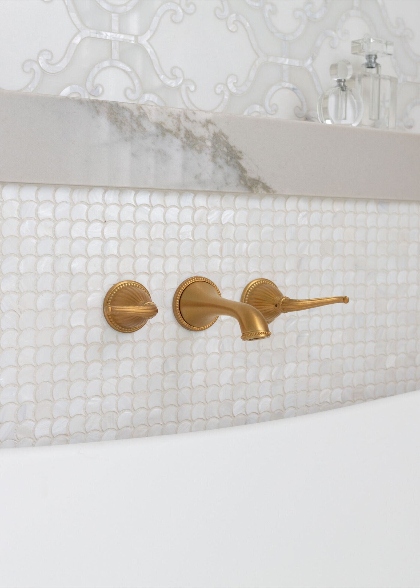 A satin brass wall-mount tub filler with fluted escutcheons and teardrop lever handles set into a wall of pearl penny-round mosaic in the Blue Heron Bathroom project by Jennifer Kizzee Design
