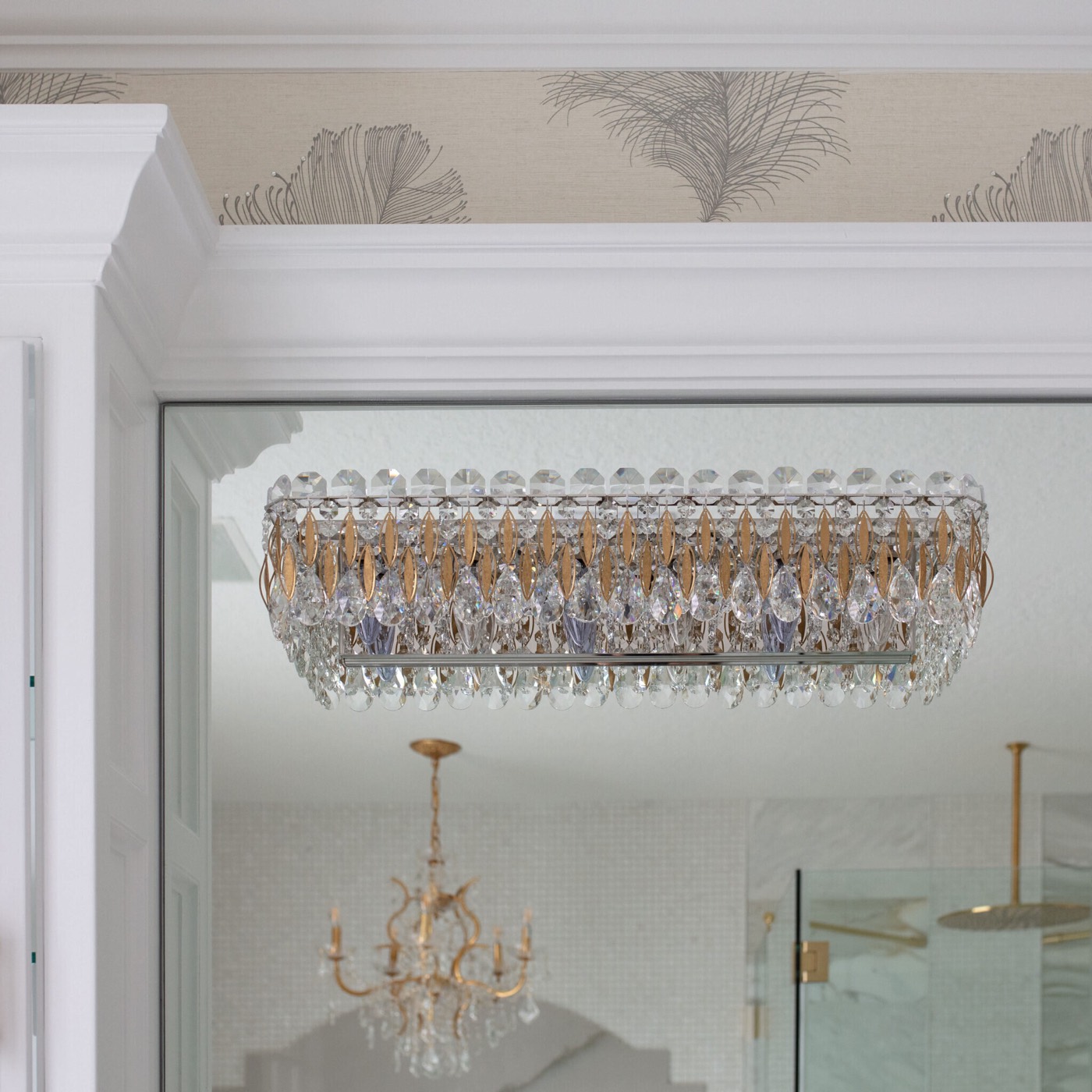 A rectangular crystal bath light with a brass frame and teardrop drops mounted directly on the mirror below white crown molding and plume-motif wallpaper in the Blue Heron Bathroom project by Jennifer Kizzee Design
