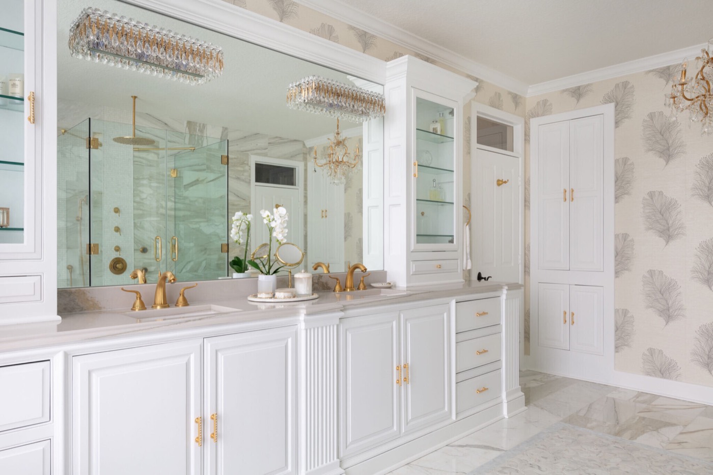 A white double vanity under a full-width mirror topped by a rectangular crystal and brass bath light, with satin brass gooseneck faucets and a glass-front cabinet tower in the Blue Heron Bathroom project by Jennifer Kizzee Design