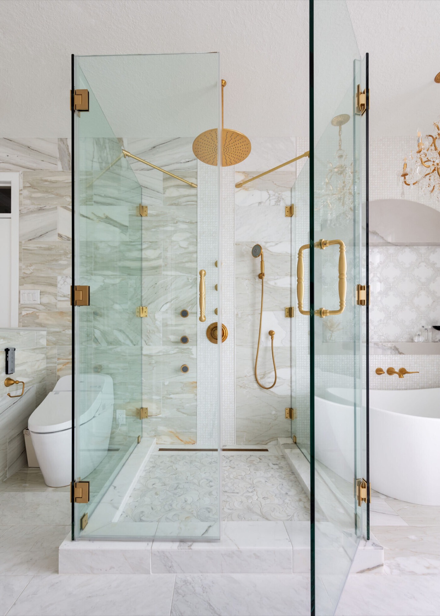 A frameless corner glass shower with satin brass hardware, marble walls and a mosaic feature stripe seen straight on beside the soaking tub in the Blue Heron Bathroom project by Jennifer Kizzee Design