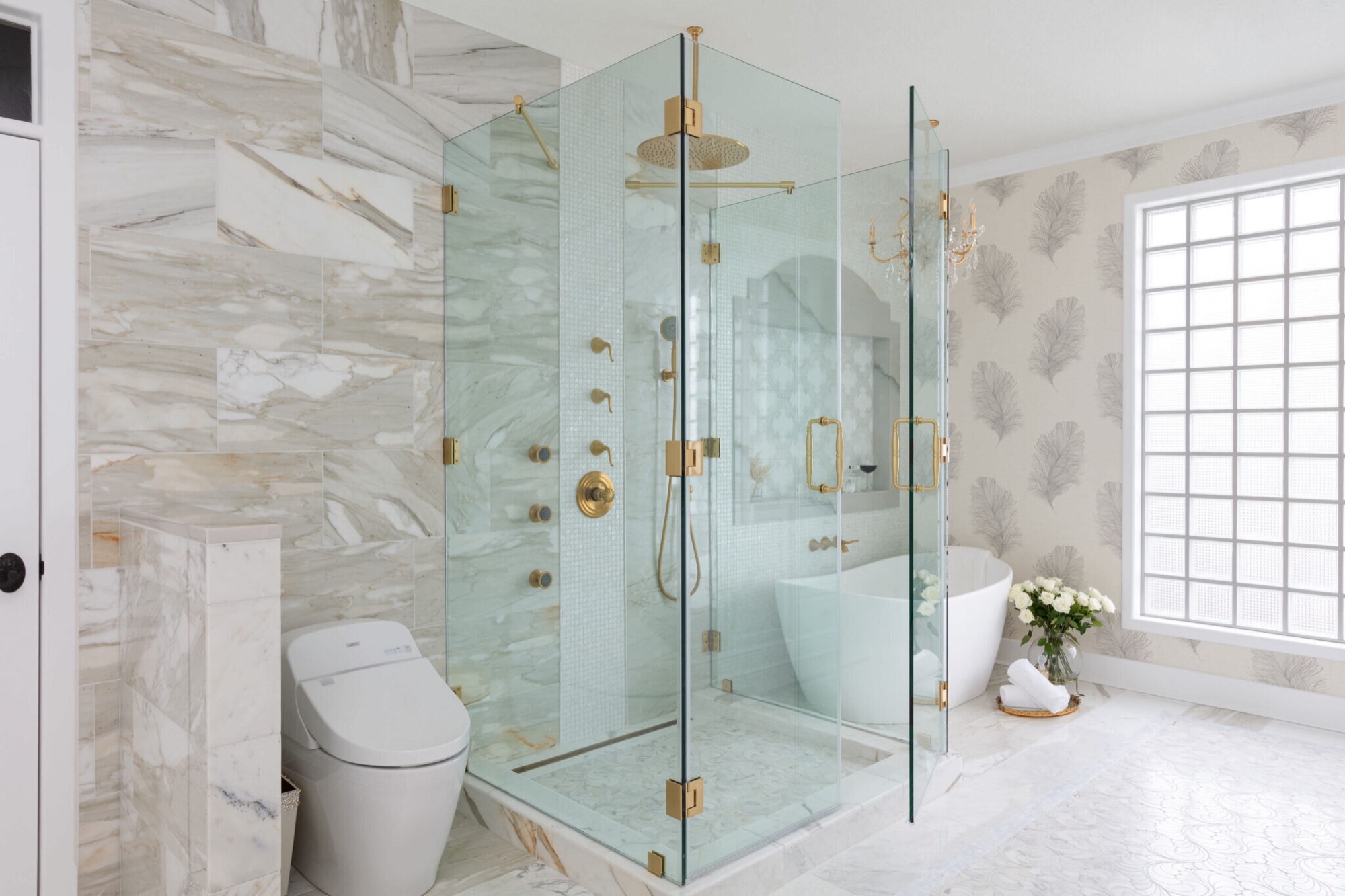 A marble-clad bathroom where a frameless corner glass shower with satin brass fixtures stands beside a freestanding soaking tub and a glass block window in the Blue Heron Bathroom project by Jennifer Kizzee Design