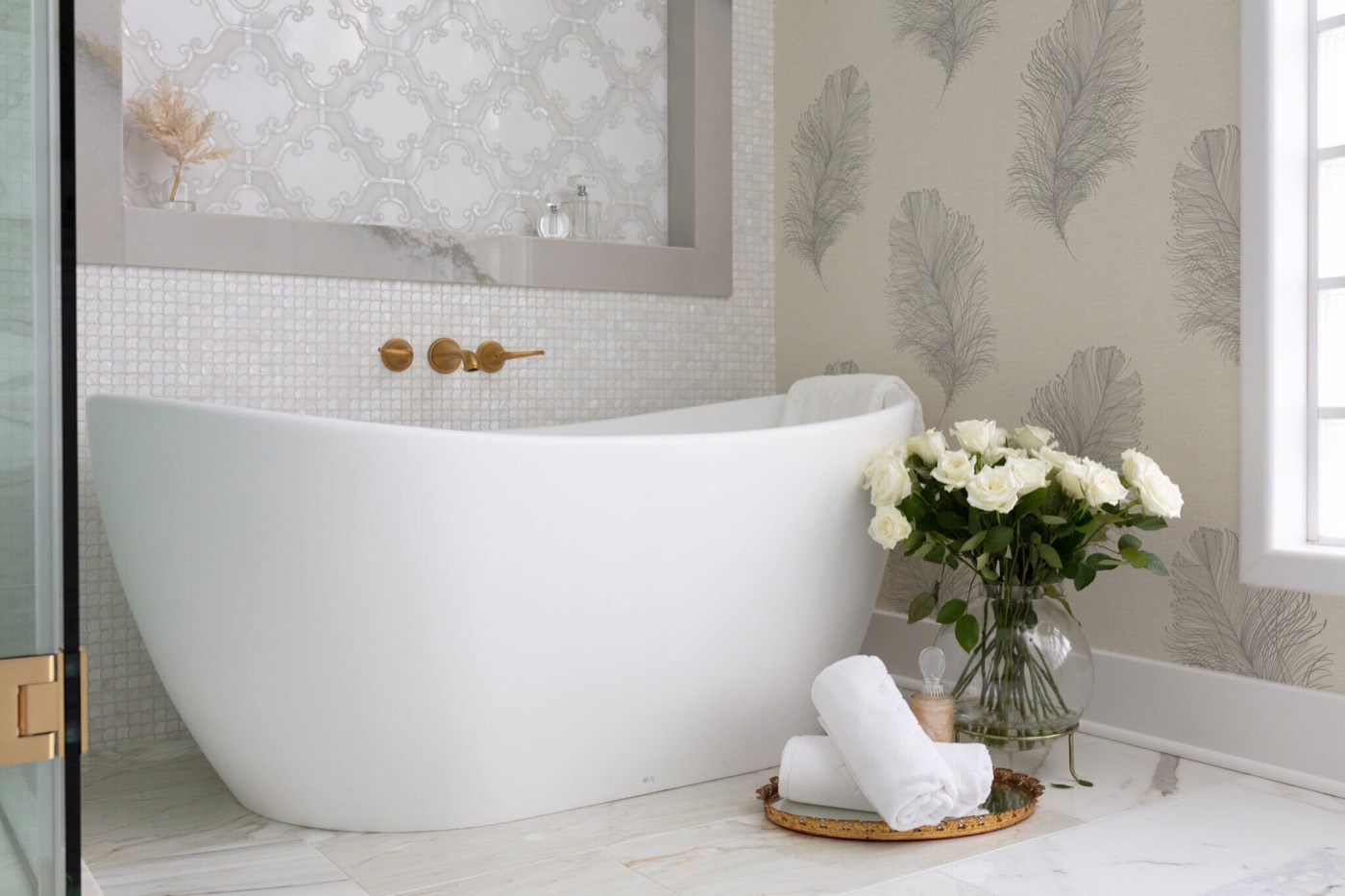 A freestanding slipper tub against a pearl penny-round mosaic wall, with a satin brass wall-mount filler and an arched tile niche above it in the Blue Heron Bathroom project by Jennifer Kizzee Design