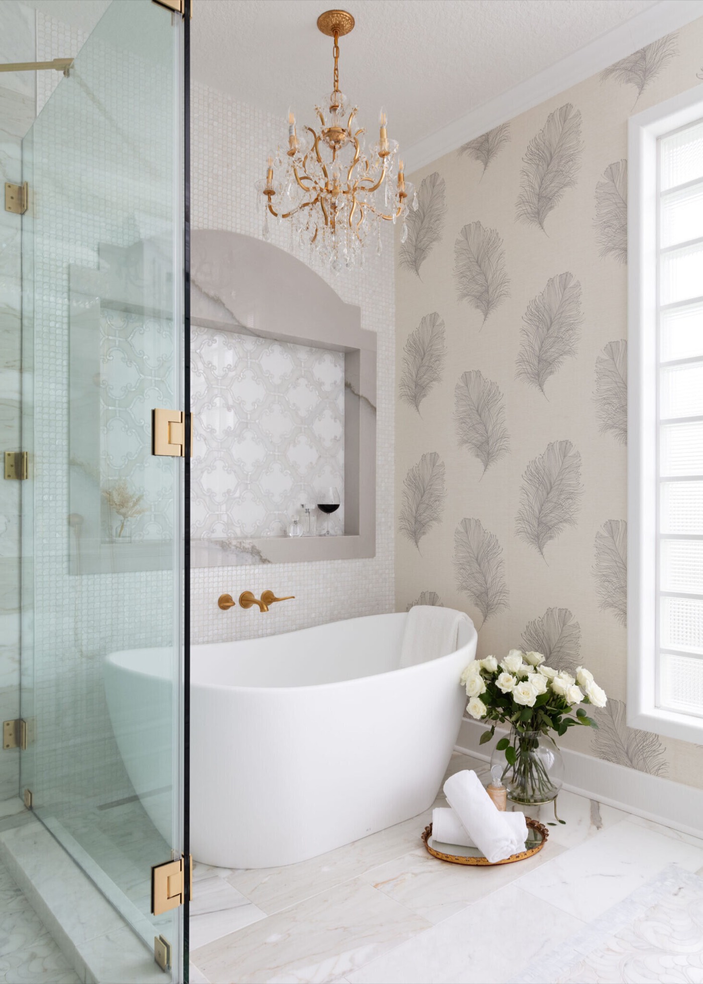 A freestanding slipper tub beneath a gold crystal chandelier, framed by pearl mosaic tile and plume-motif wallpaper in the Blue Heron Bathroom project by Jennifer Kizzee Design