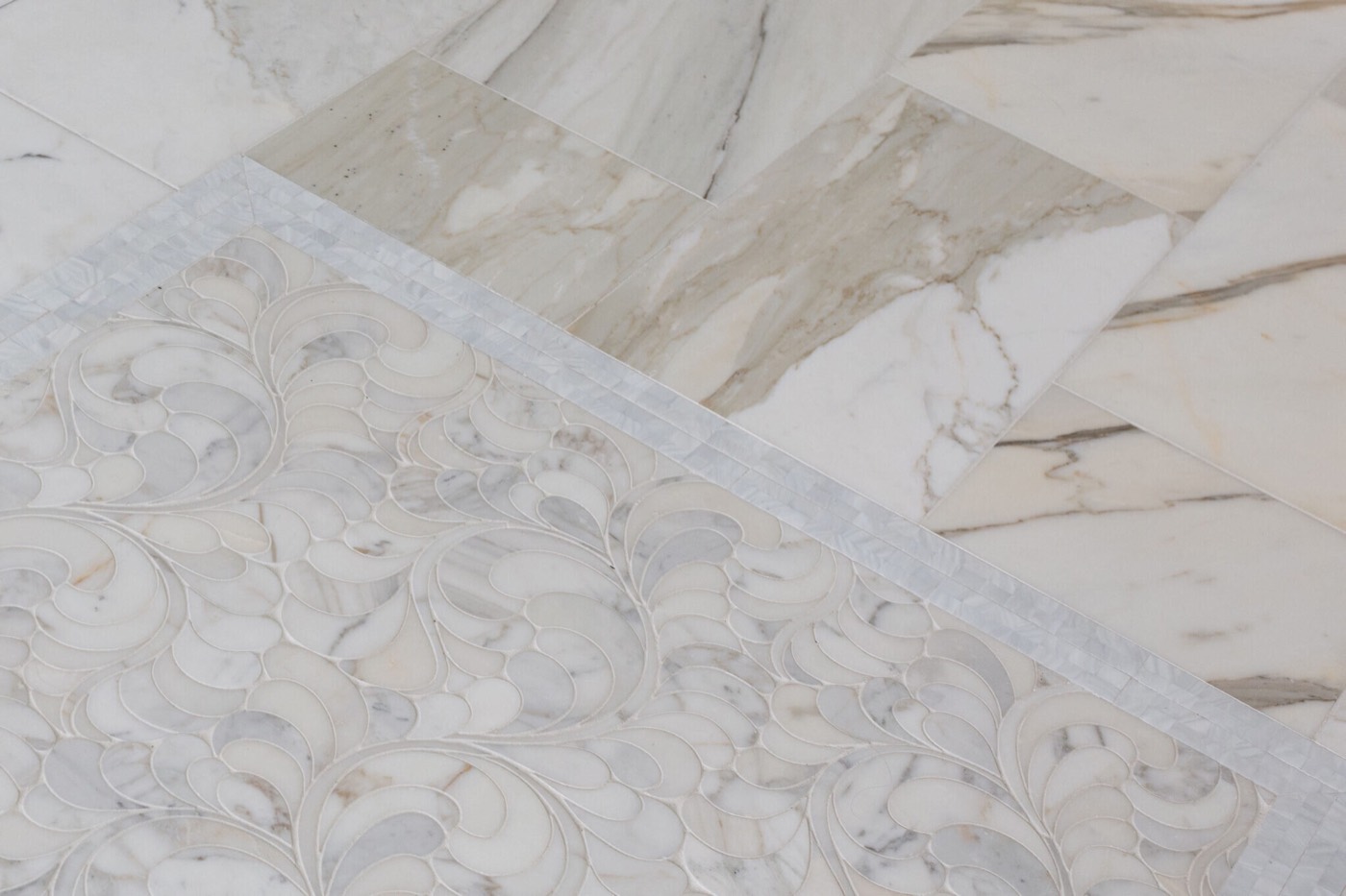 A marble floor where a waterjet mosaic panel in a swirling plume pattern meets large marble tiles across a narrow mother-of-pearl border in the Blue Heron Bathroom project by Jennifer Kizzee Design