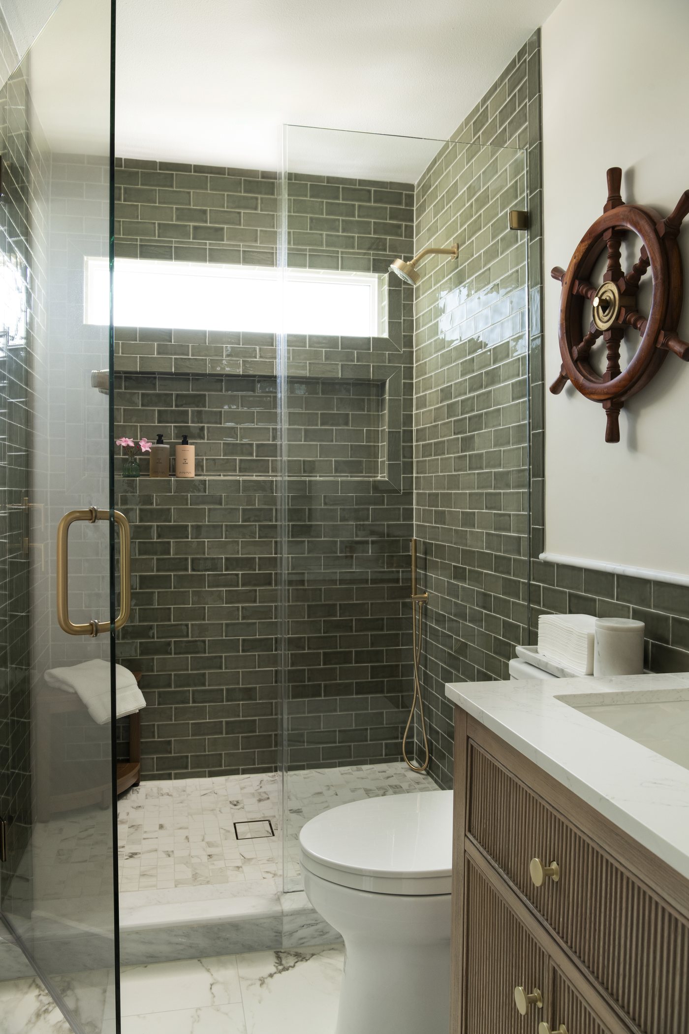 Shower in green tile with a glass door and a ship's wheel on the wall in the Bolivar Beach House project by Jennifer Kizzee Design