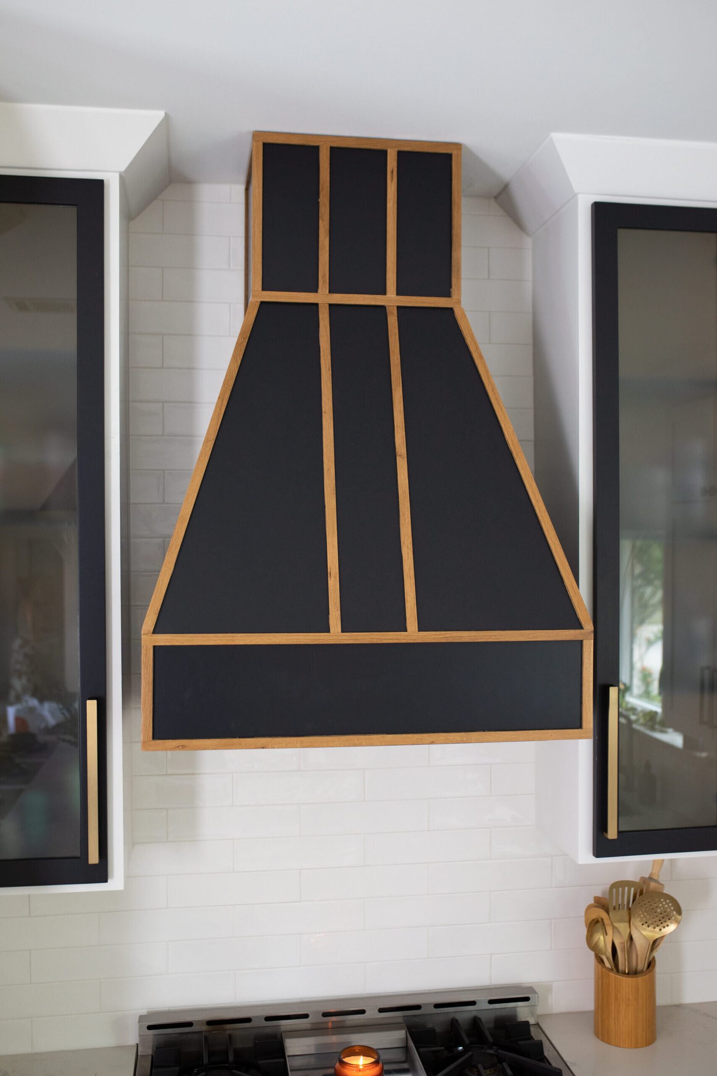 Black range hood banded in brass above a range, flanked by black glass front cabinets in the Branstetter Kitchen project by Jennifer Kizzee Design