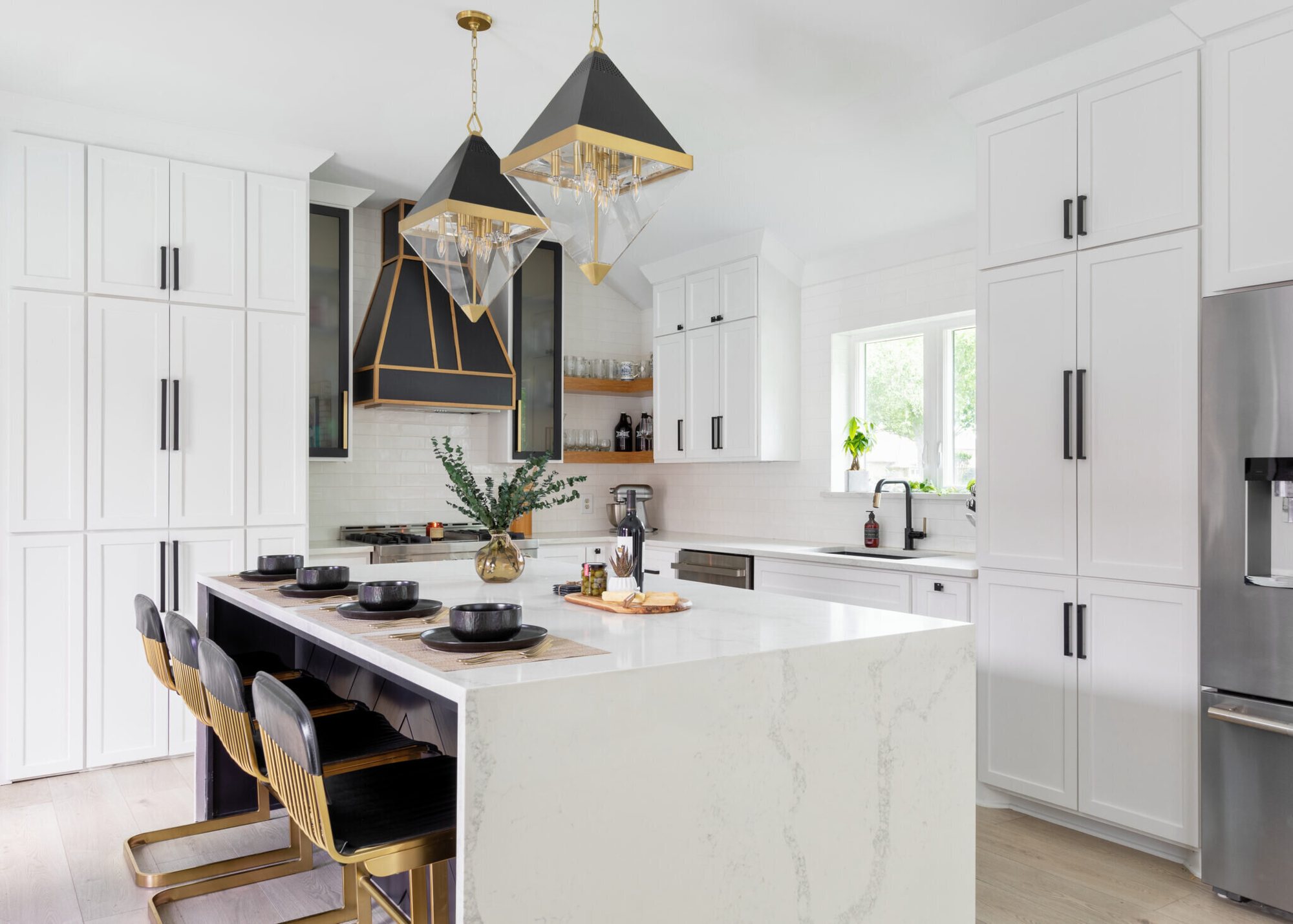 White kitchen with a black and brass range hood, a long quartz island and faceted pendants in the Branstetter Kitchen project by Jennifer Kizzee Design