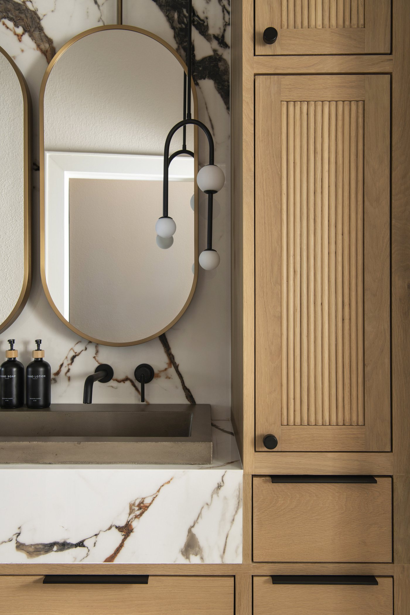 Oval mirror against a marble slab wall beside fluted white oak storage in the Collective Washroom project by Jennifer Kizzee Design
