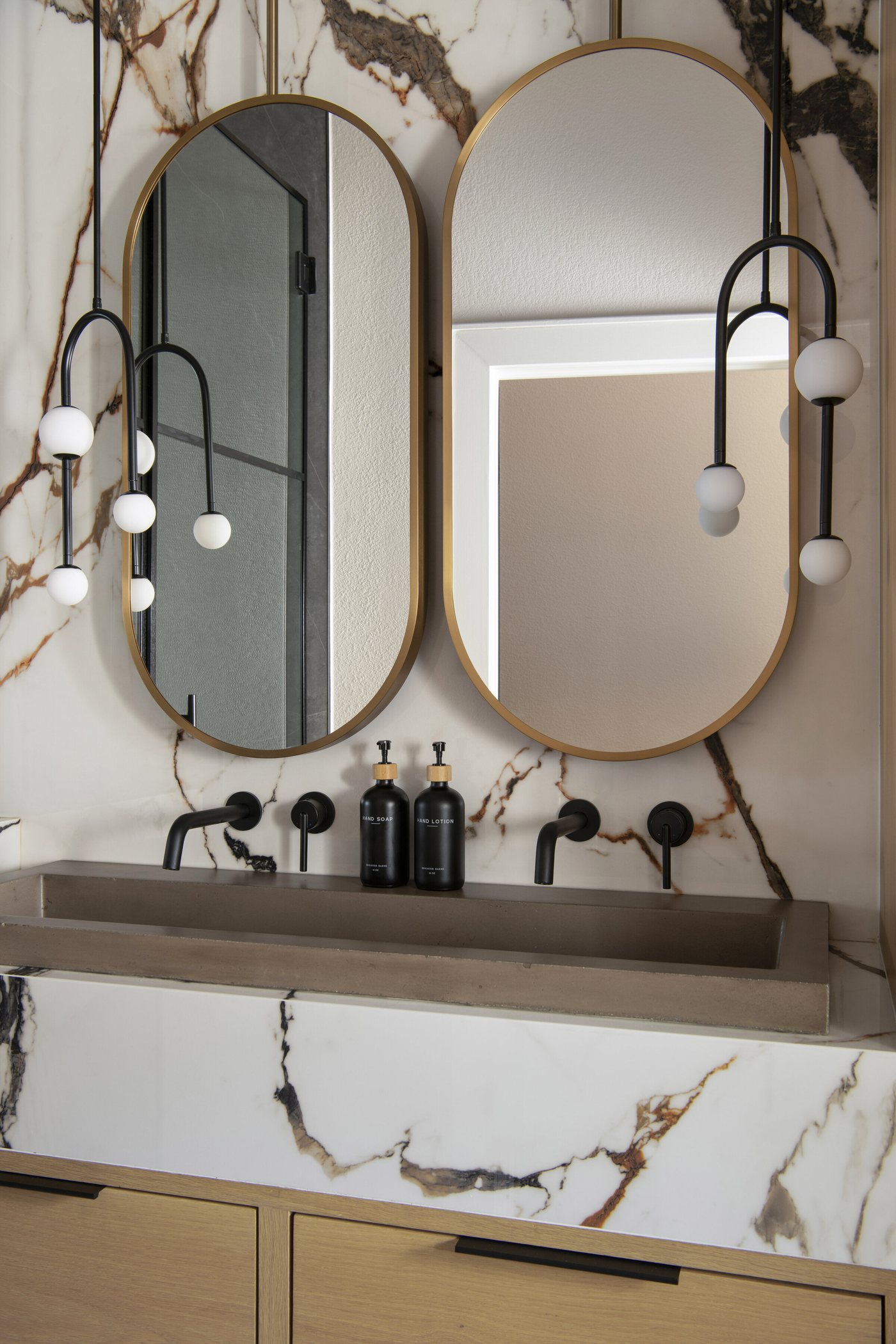 Twin oval mirrors with wall mounted black faucets over a stone trough in the Collective Washroom project by Jennifer Kizzee Design