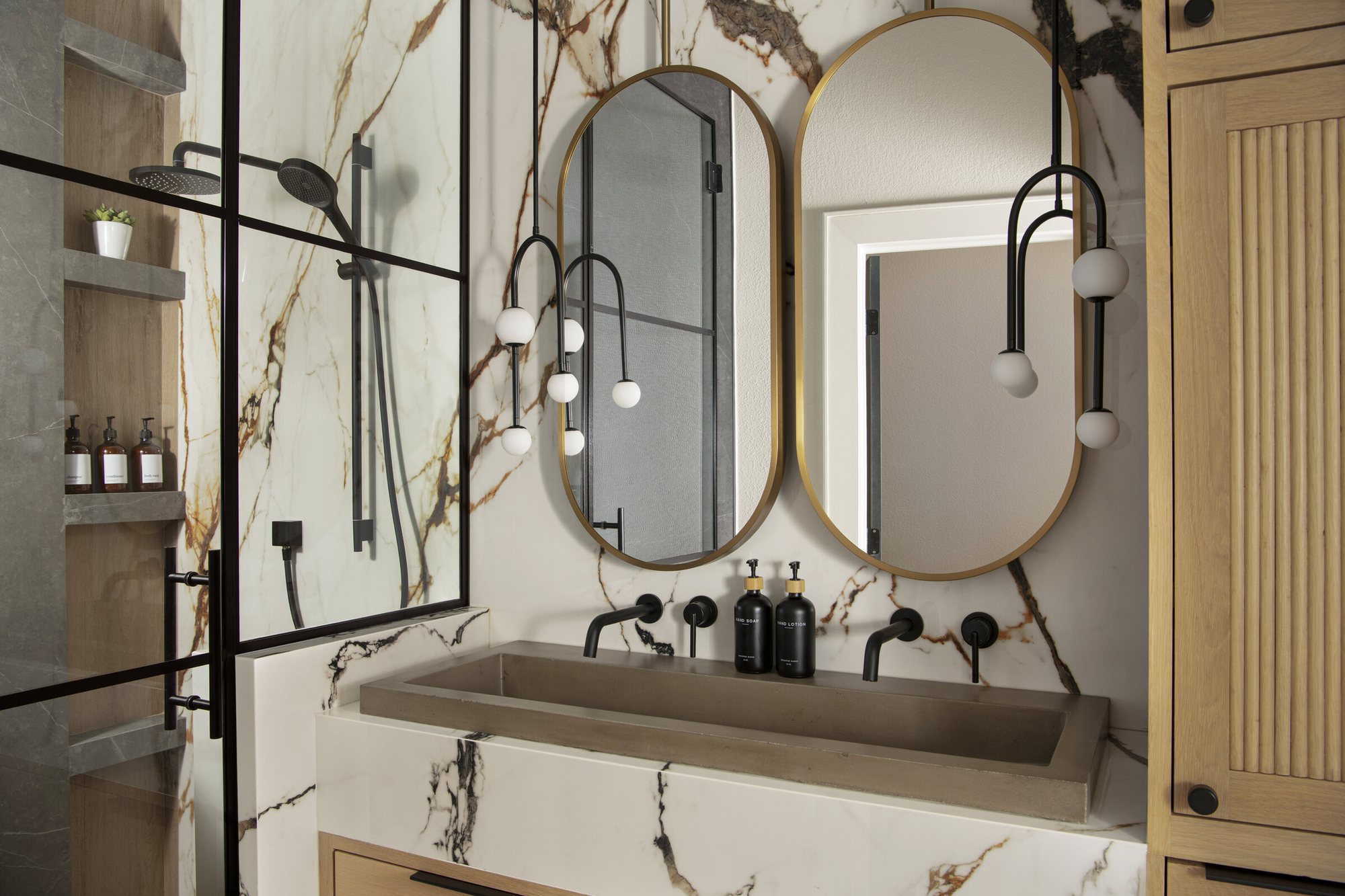 Washroom with a marble slab wall, twin oval mirrors and a stone trough sink in the Collective Washroom project by Jennifer Kizzee Design