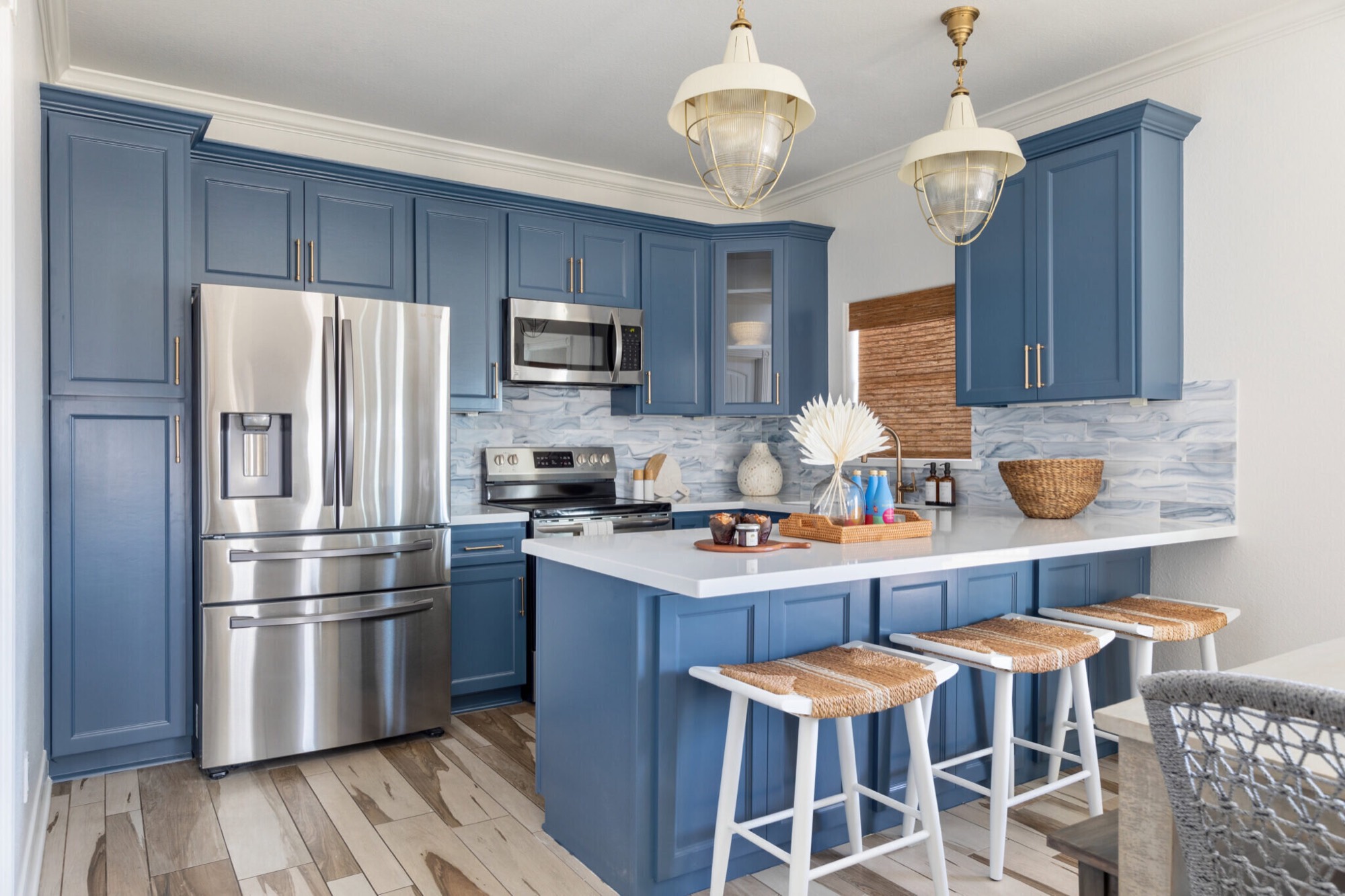 Kitchen in denim blue shaker cabinetry with glass and brass pendants and woven seagrass counter stools in the Crystal Beach Vacation Home project by Jennifer Kizzee Design