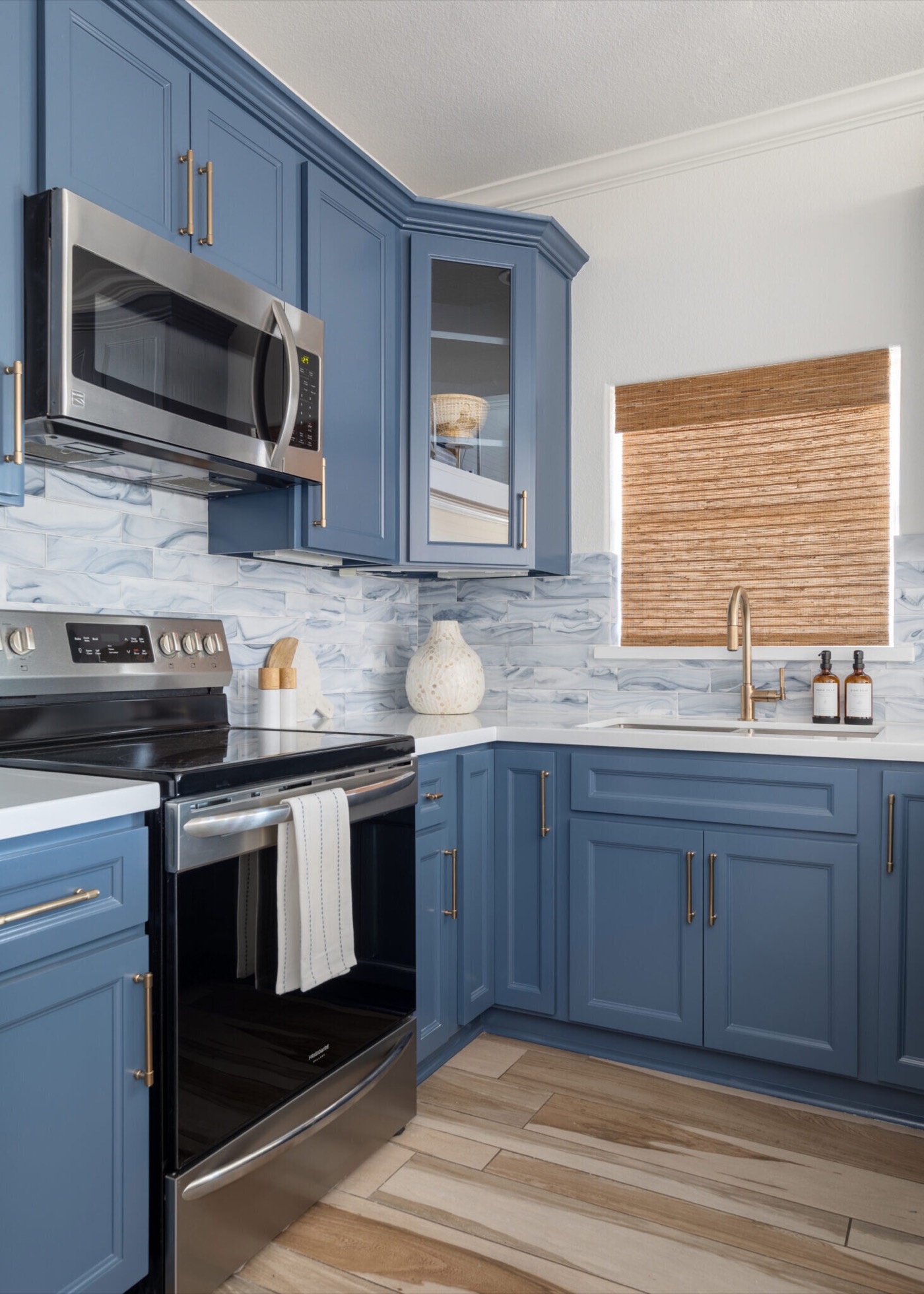 Range corner with a marble-look tile backsplash, a brass faucet, and a woven wood shade in the Crystal Beach Vacation Home project by Jennifer Kizzee Design
