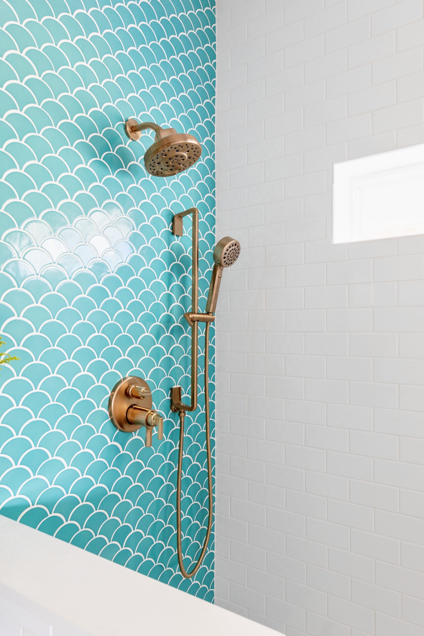 Brass shower head and hand shower where turquoise fish-scale tile meets white subway tile in the Crystal Beach Vacation Home project by Jennifer Kizzee Design