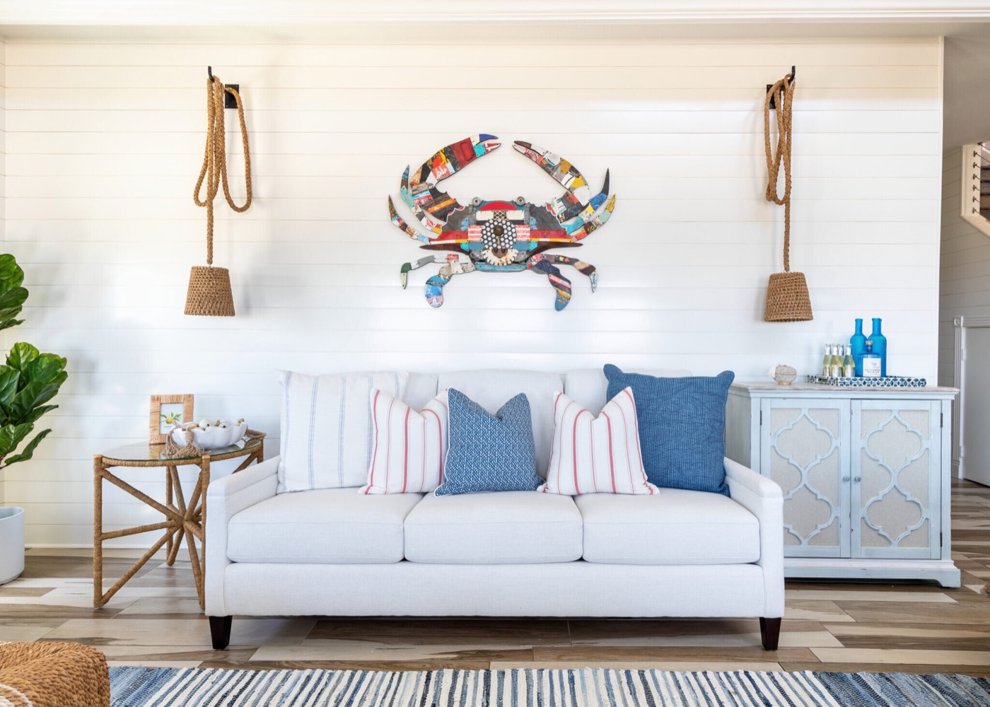 White sofa on a shiplap wall beneath a multicolor crab sculpture flanked by rope-hung woven pendants in the Crystal Beach Vacation Home project by Jennifer Kizzee Design