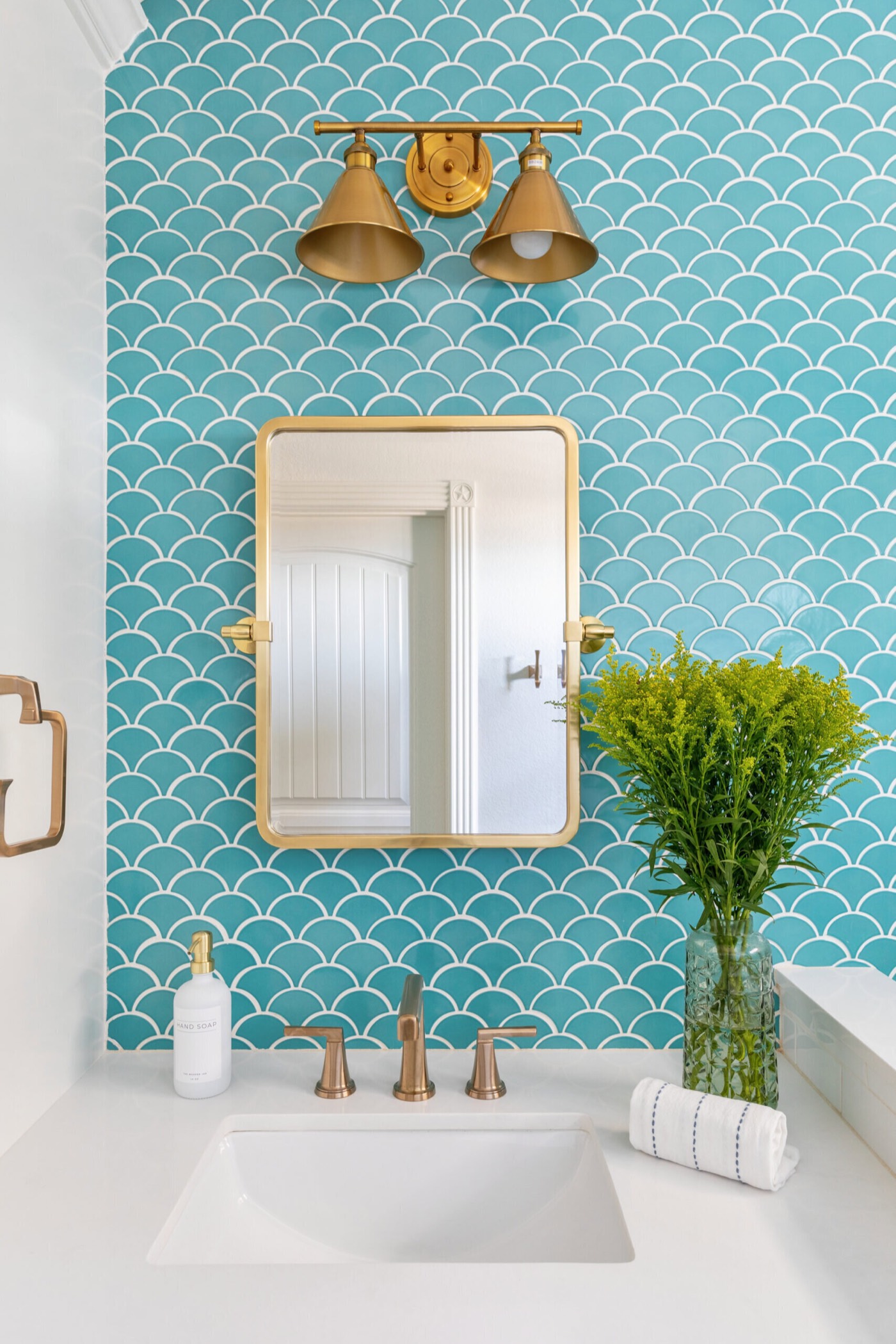 Brass two-light sconce and gold pivot mirror set against a turquoise fish-scale tile wall in the Crystal Beach Vacation Home project by Jennifer Kizzee Design