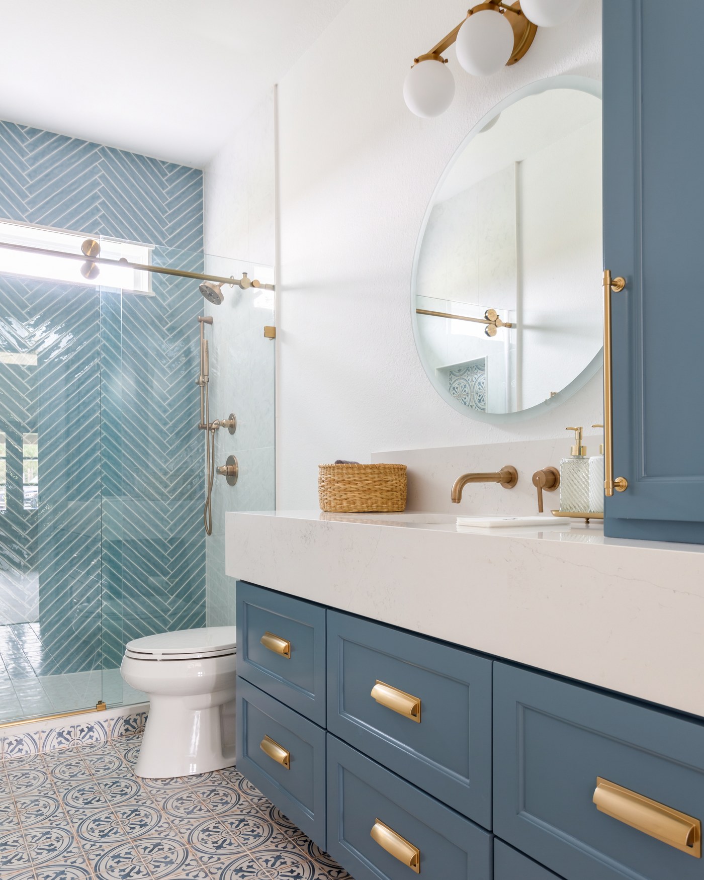 Bathroom with a steel blue vanity, a round mirror, and a walk-in shower in light blue herringbone tile in the Emma Lane Guest House project by Jennifer Kizzee Design