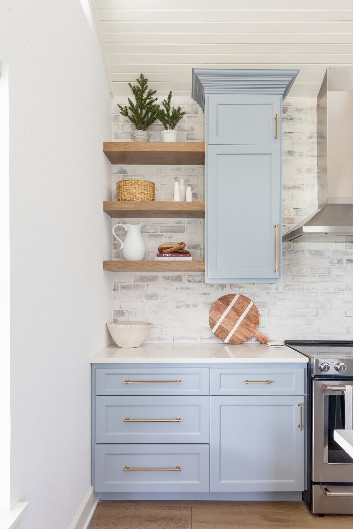 Oak floating shelves mounted on a whitewashed brick backsplash beside a powder blue upper cabinet in the Emma Lane Guest House project by Jennifer Kizzee Design