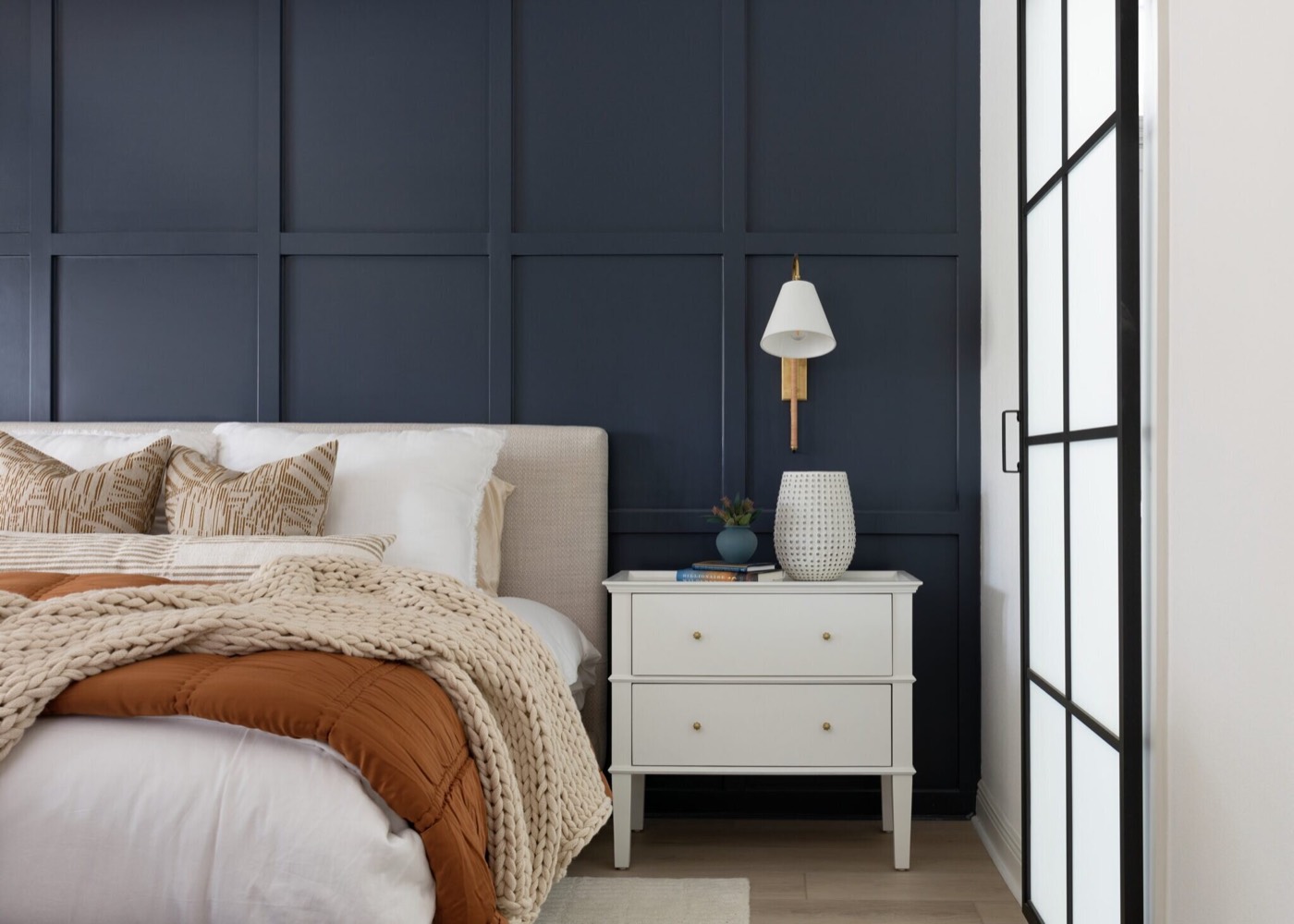 Oatmeal upholstered headboard and white two-drawer nightstand against the navy grid panelled wall in the Evergreen Suite Bedroom project by Jennifer Kizzee Design
