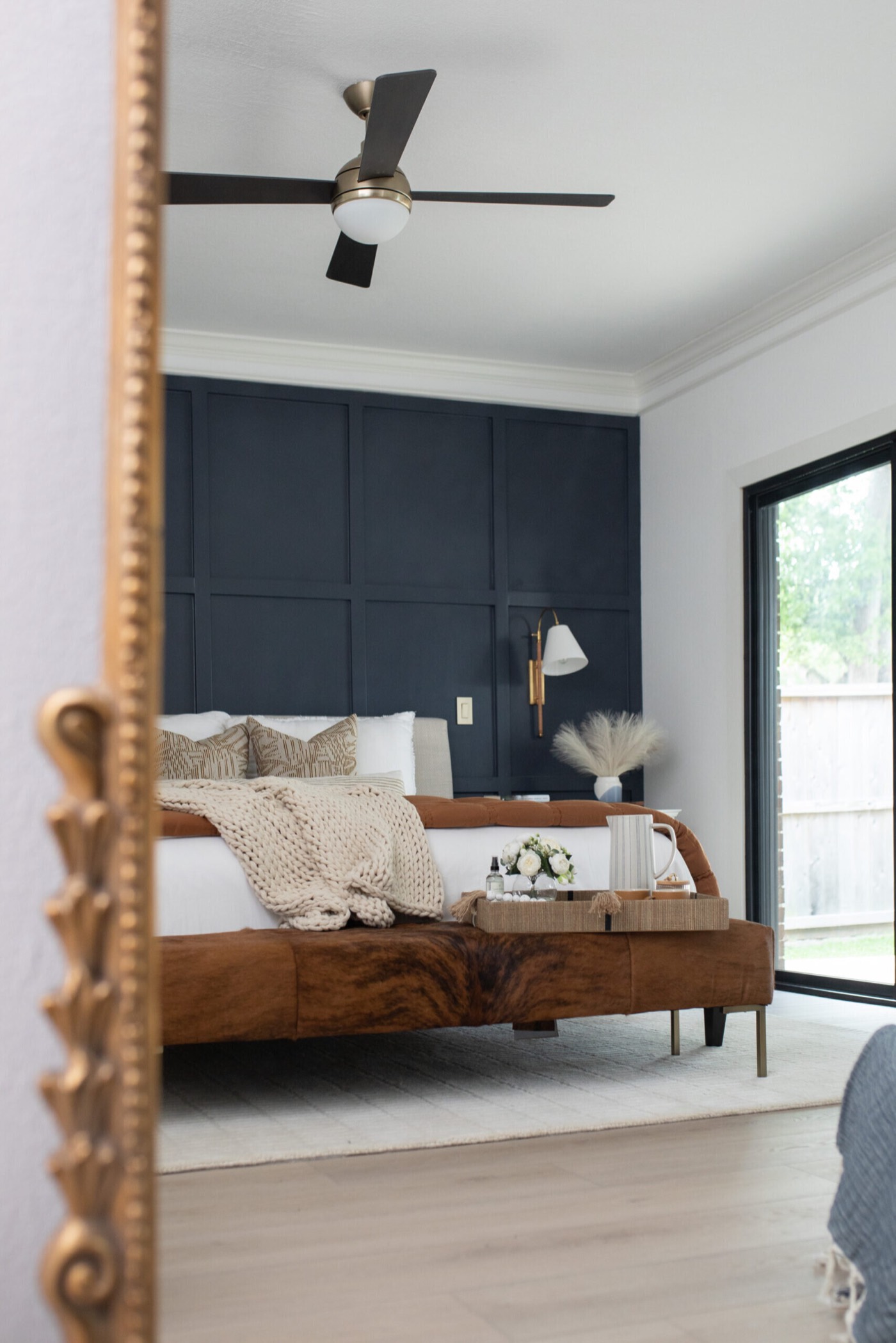 The bed and navy panelled wall seen past the carved gold mirror frame in the Evergreen Suite Bedroom project by Jennifer Kizzee Design