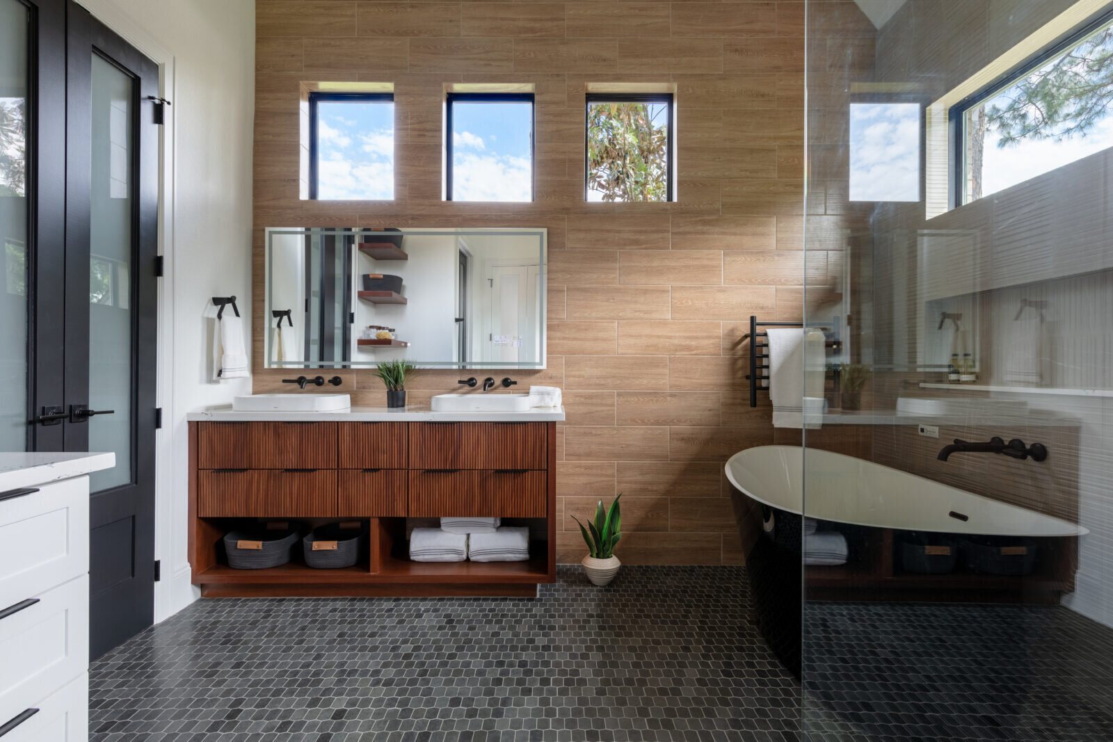 Modern primary bath with a fluted walnut vanity, black freestanding tub and black hex mosaic floor in the Fox Run Modern Bath project by Jennifer Kizzee Design