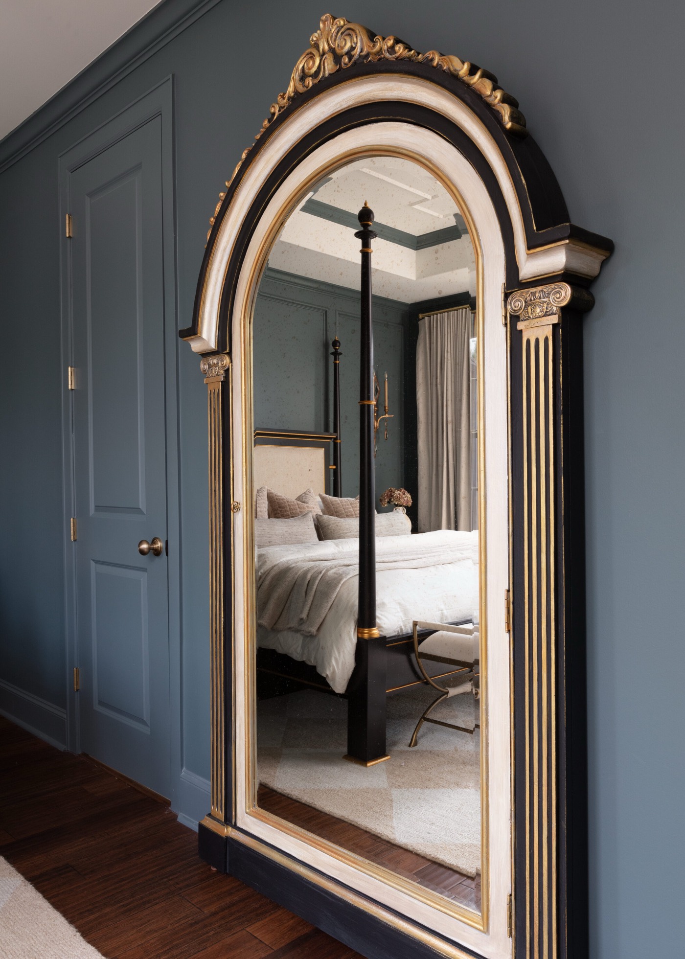Gilt and black arched floor mirror reflecting the four-poster bed against a dark teal wall in the Gateway Condo project by Jennifer Kizzee Design