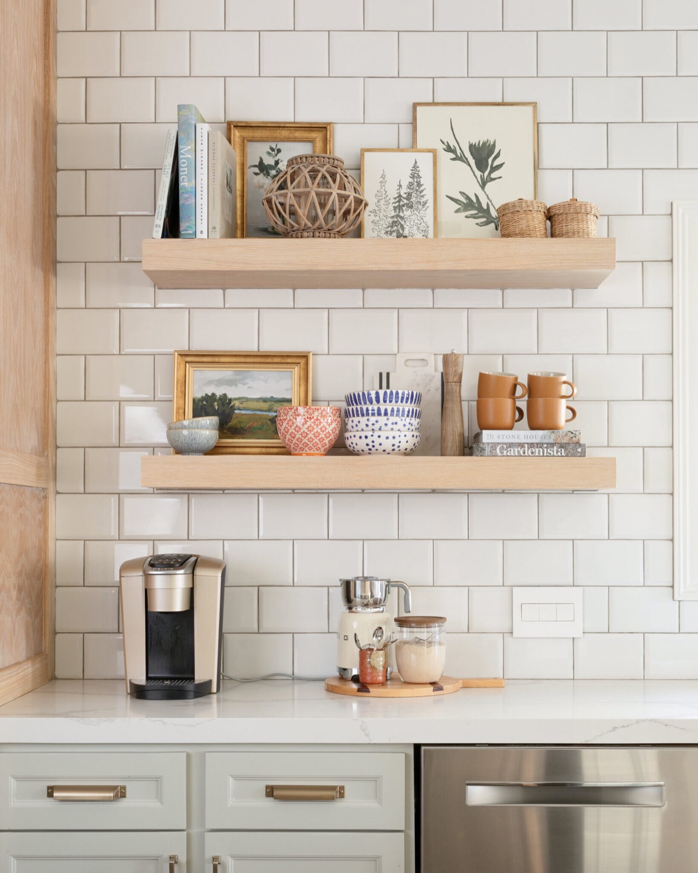 Coffee station tucked beneath two white oak floating shelves on white tile beside the stainless dishwasher in the Jenn's Kitchen project by Jennifer Kizzee Design