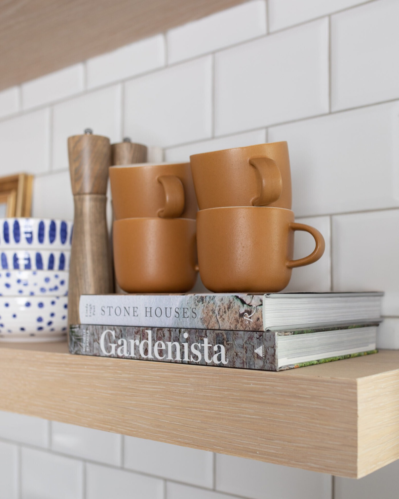 Stacked terracotta mugs and a wood pepper mill on a white oak floating shelf against white handmade tile in the Jenn's Kitchen project by Jennifer Kizzee Design