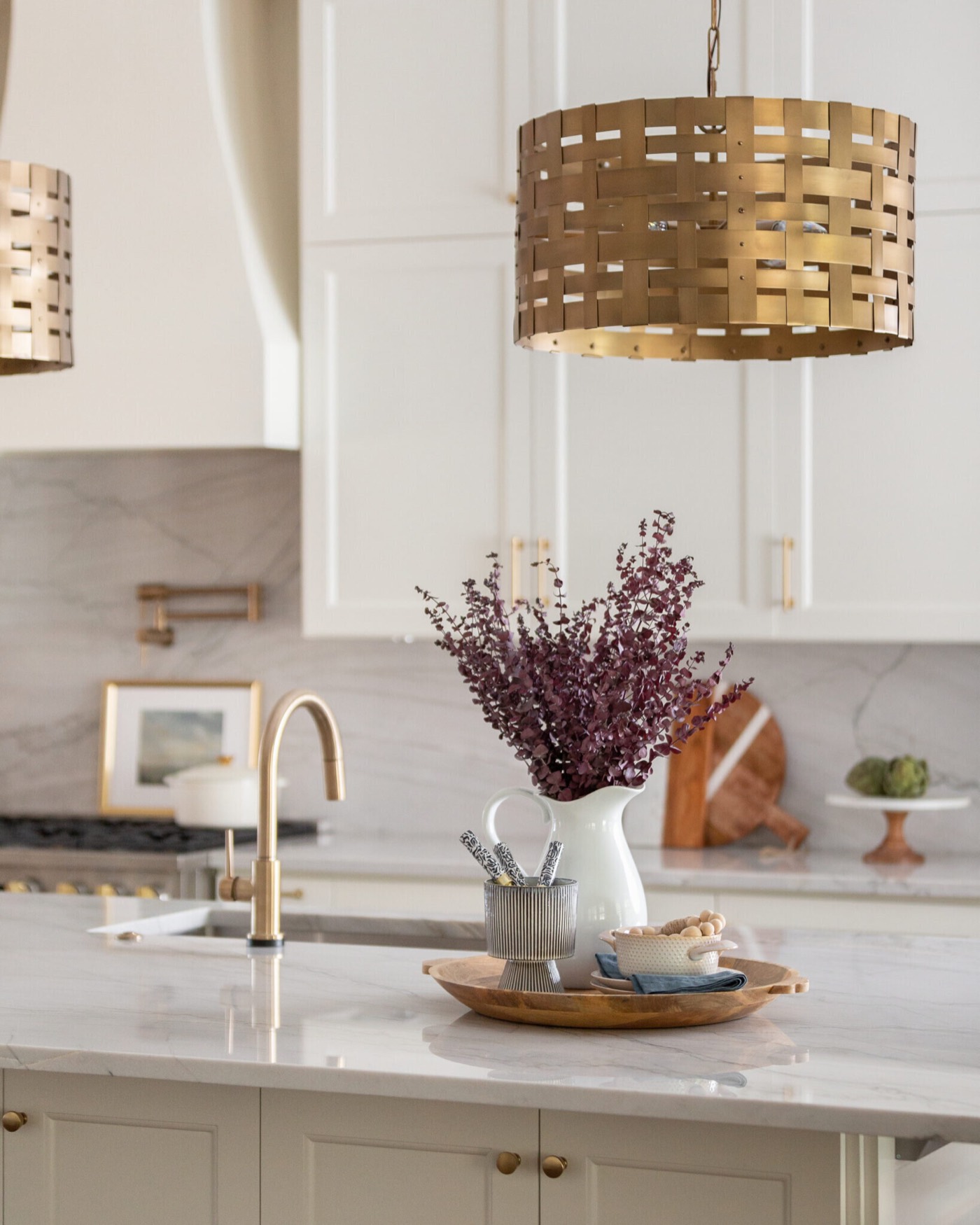 Brass gooseneck faucet and a white pitcher of heather on the island stone beneath a woven brass drum pendant in the Knoll Kitchen project by Jennifer Kizzee Design