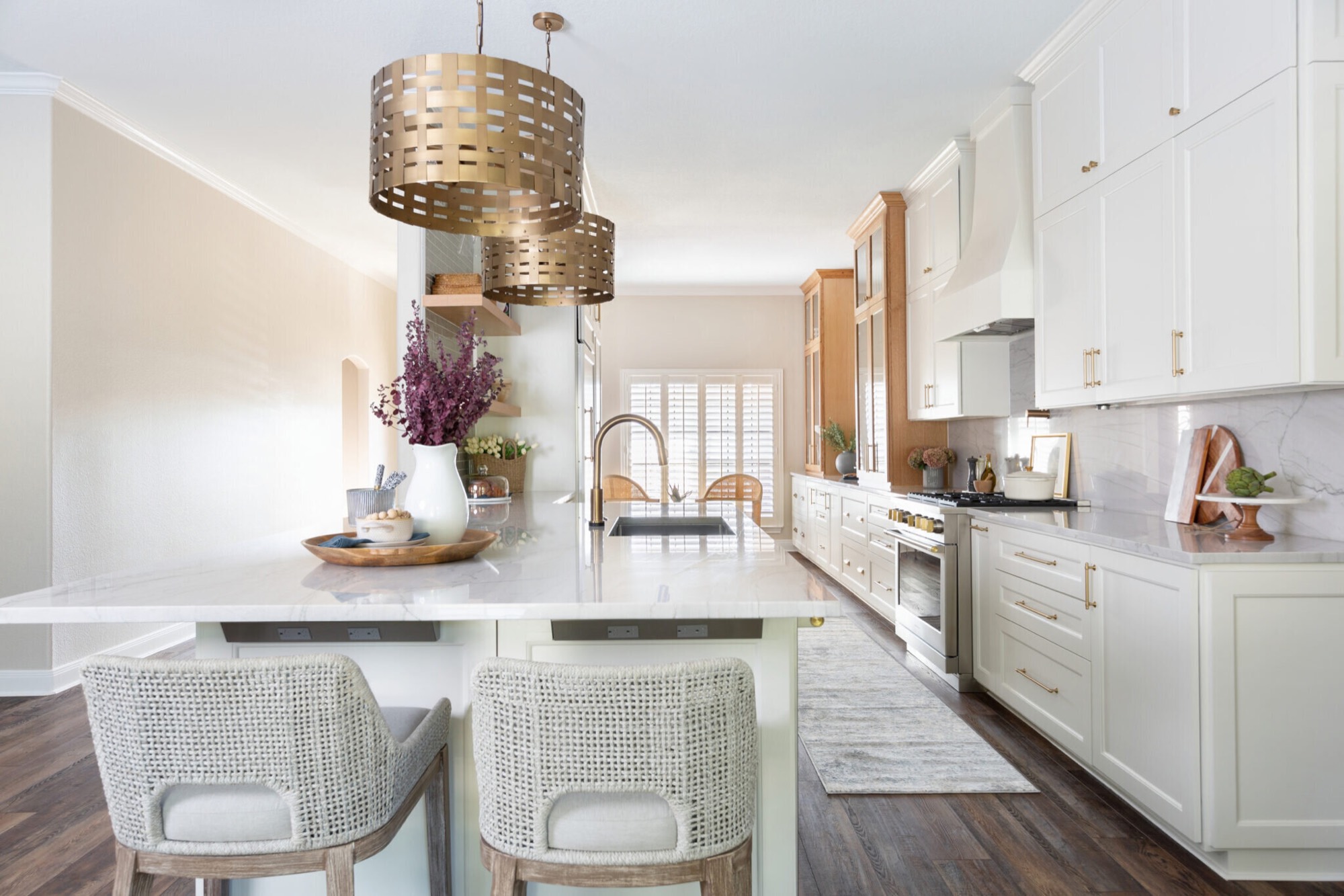 Kitchen island in white veined stone beneath two woven brass drum pendants, with the plaster range hood and marble slab backsplash beyond, in the Knoll Kitchen project by Jennifer Kizzee Design