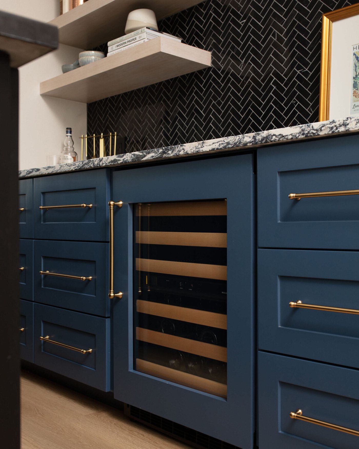 Navy cabinetry with brass pulls and a glass front wine refrigerator in the Markee Oaks project by Jennifer Kizzee Design