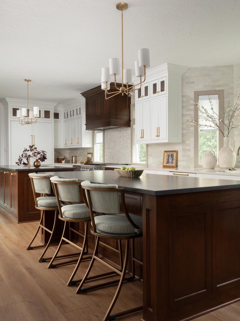 Dark stained island with green leather counter stools under brass chandeliers in the Pebble Brook project by Jennifer Kizzee Design