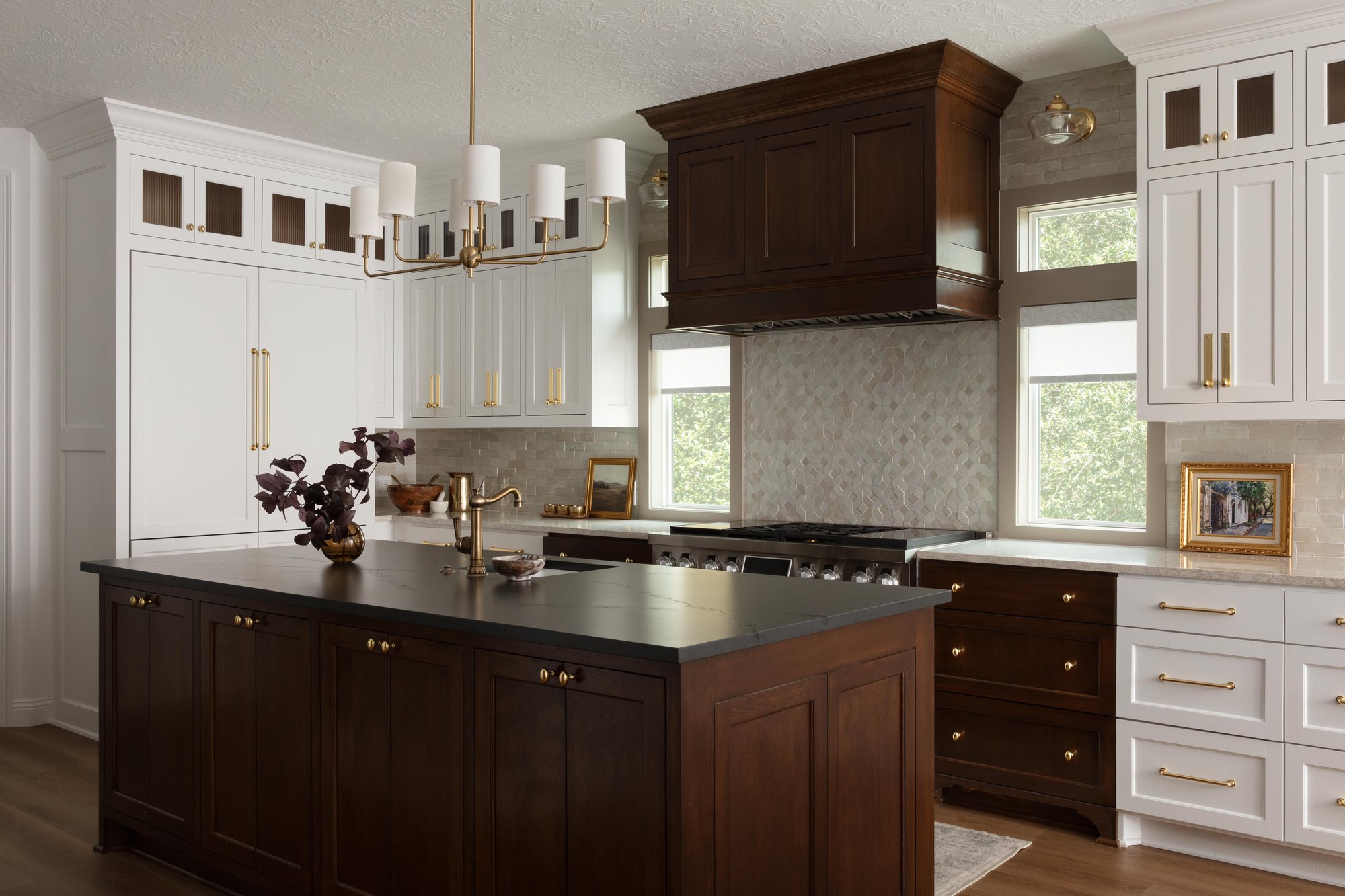 Kitchen with dark stained cabinetry, a wood hood and a marble backsplash under brass chandeliers in the Pebble Brook project by Jennifer Kizzee Design