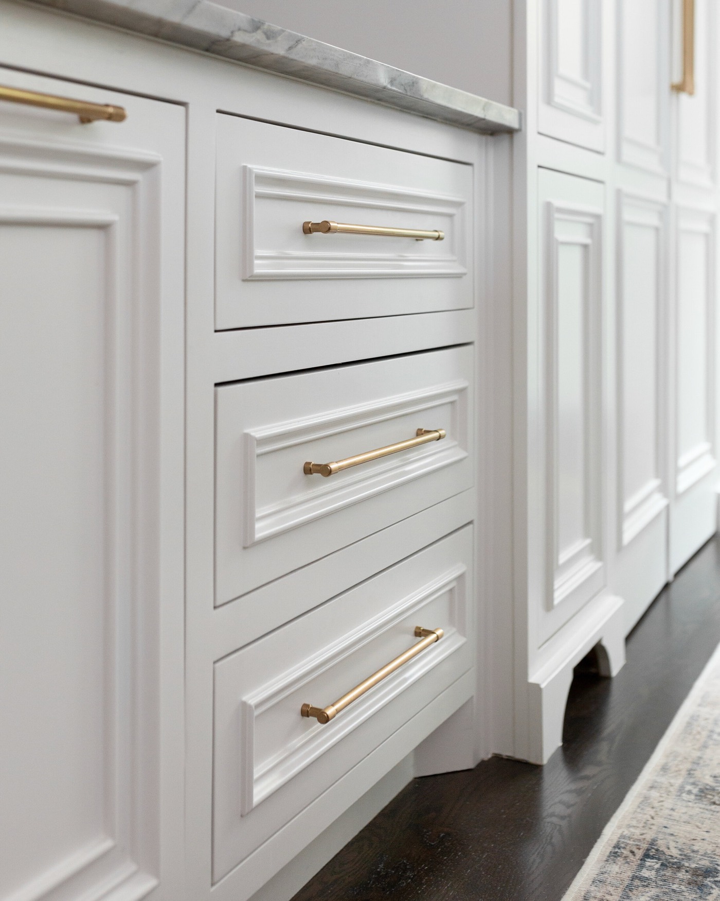 Aged brass bar pulls on the white raised panel drawer stack below the veined stone counter in the Pinewold Kitchen project by Jennifer Kizzee Design