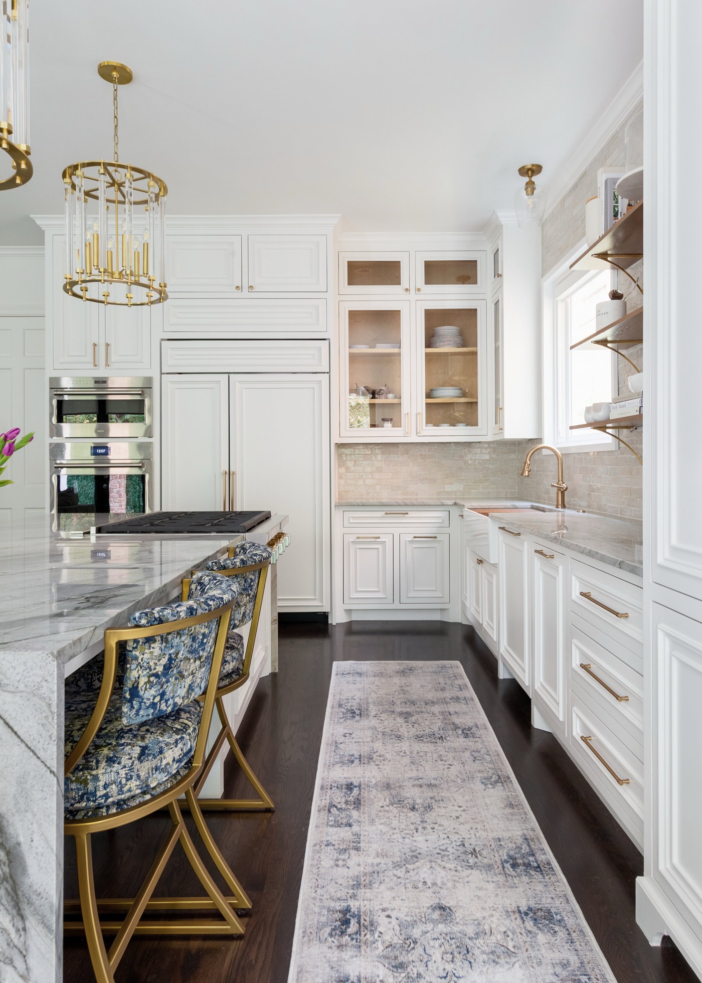 View along the kitchen from the island's blue patterned stools to the brass faucet and apron sink at the window in the Pinewold Kitchen project by Jennifer Kizzee Design