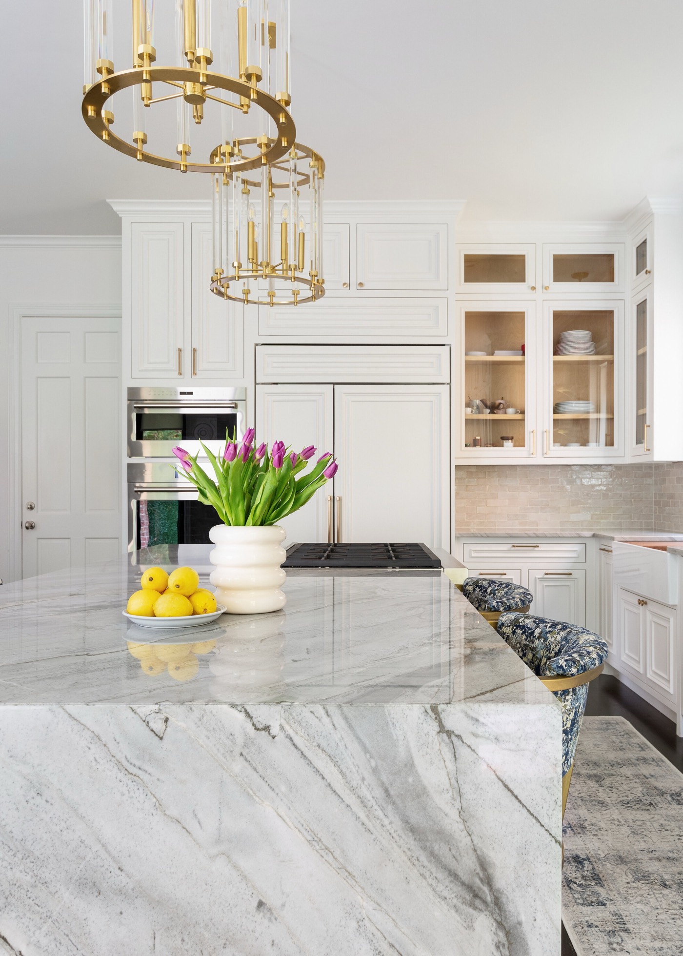 Heavily veined gray and white stone island top styled with tulips and lemons beneath two brass lantern pendants in the Pinewold Kitchen project by Jennifer Kizzee Design