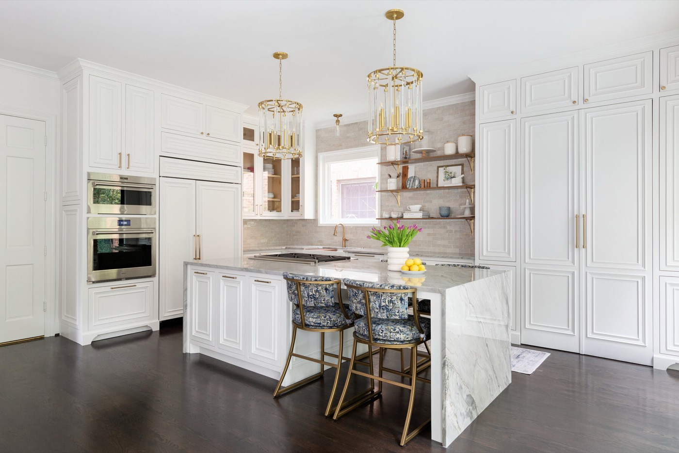 Paneled wall ovens beside a veined stone island under glass and brass lantern pendants in the Pinewold Kitchen project by Jennifer Kizzee Design