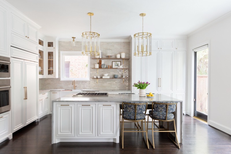 White raised panel kitchen with a veined stone waterfall island under two brass and glass lantern pendants in the Pinewold Kitchen project by Jennifer Kizzee Design