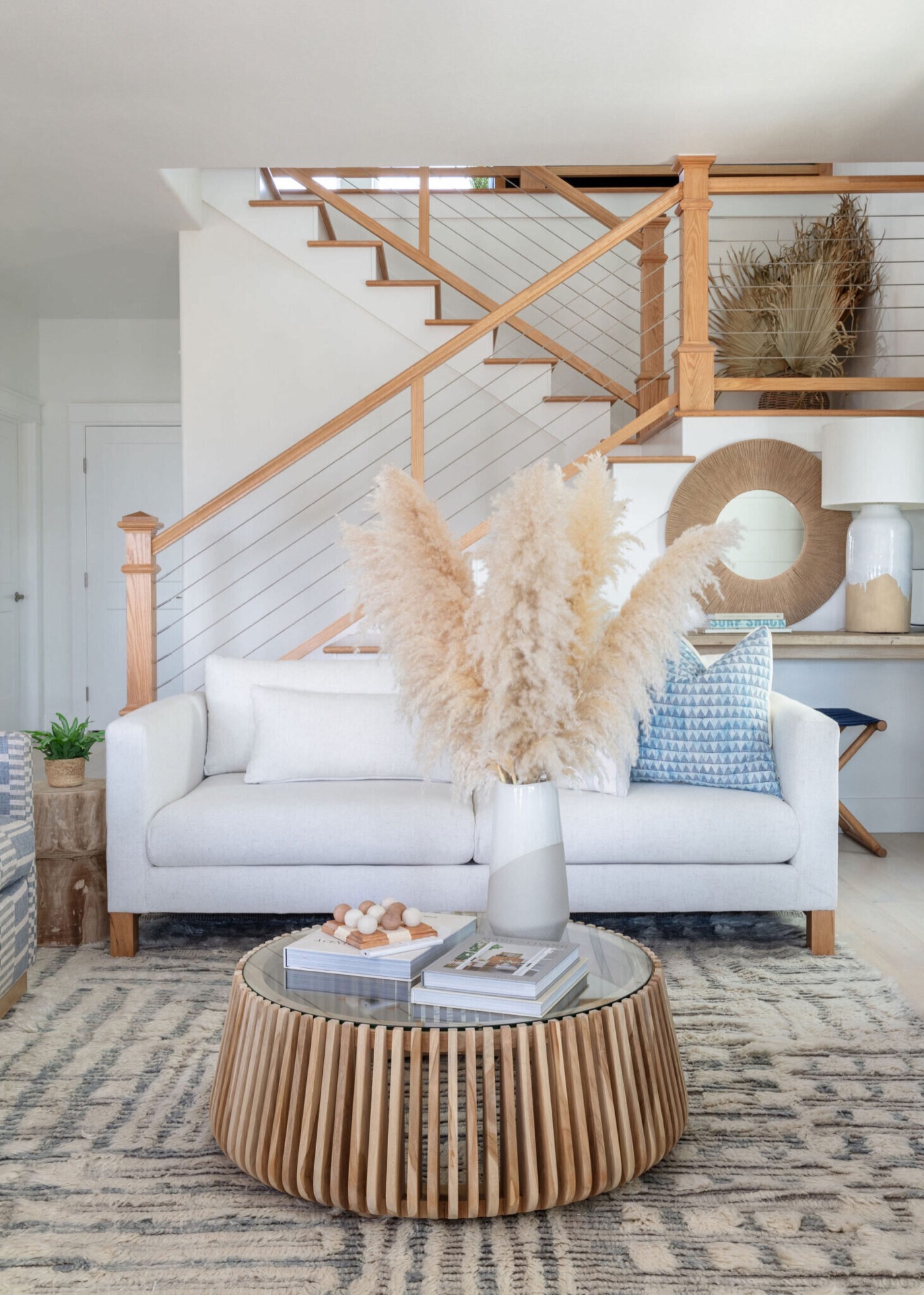 White sofa beneath an oak and cable-rail staircase with pampas grass on a slatted round coffee table in the Starfish Vacation Home project by Jennifer Kizzee Design