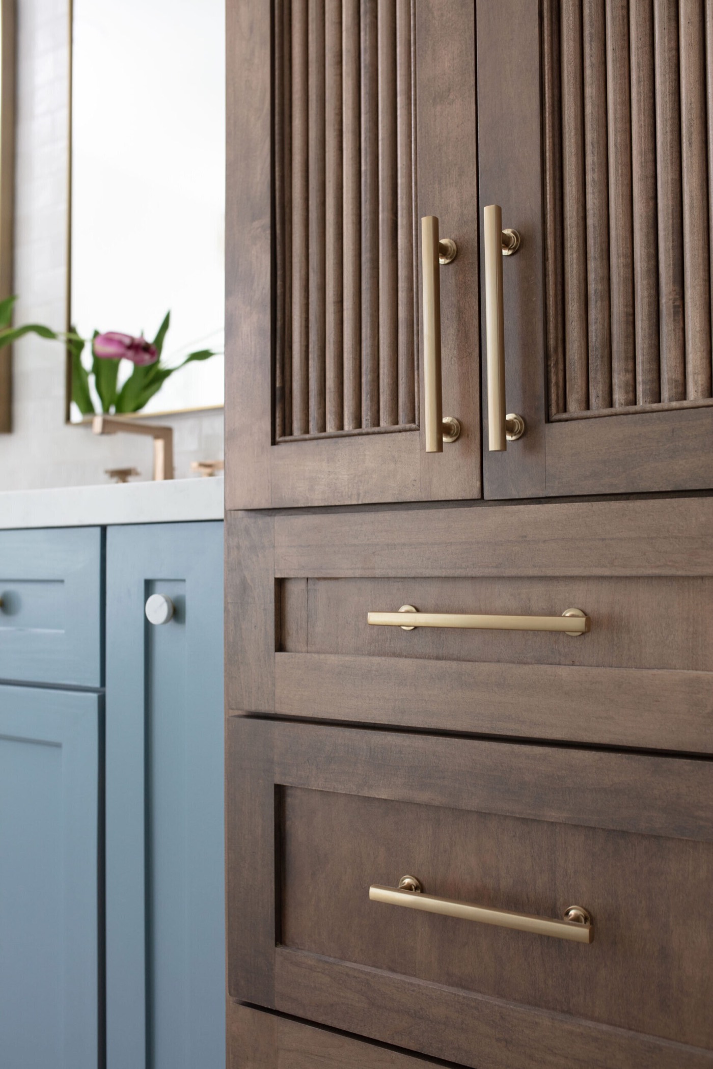 Brass bar pulls on a reeded walnut linen tower next to the dusty blue vanity in the Sunstream Bath project by Jennifer Kizzee Design