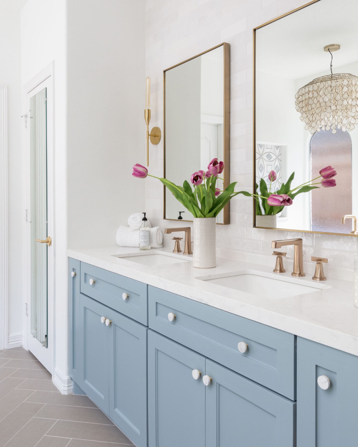 Dusty blue vanity with tulips, brass mirrors and the capiz chandelier reflected in the glass in the Sunstream Bath project by Jennifer Kizzee Design