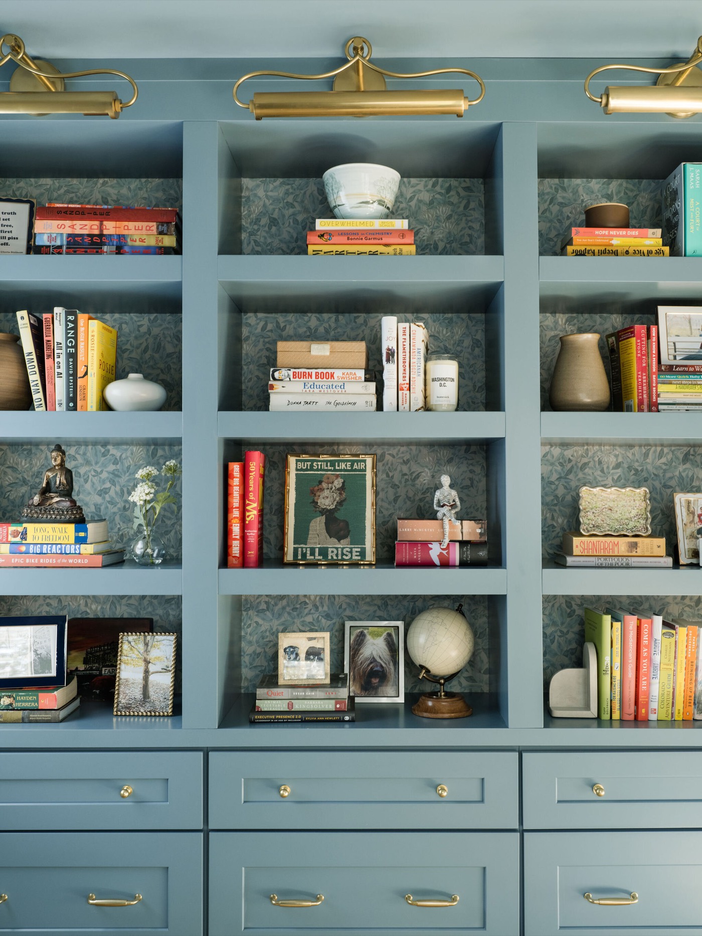 Blue painted library built ins backed in blue green texture and lit by three brass picture lights in the Takoma Park project by Jennifer Kizzee Design