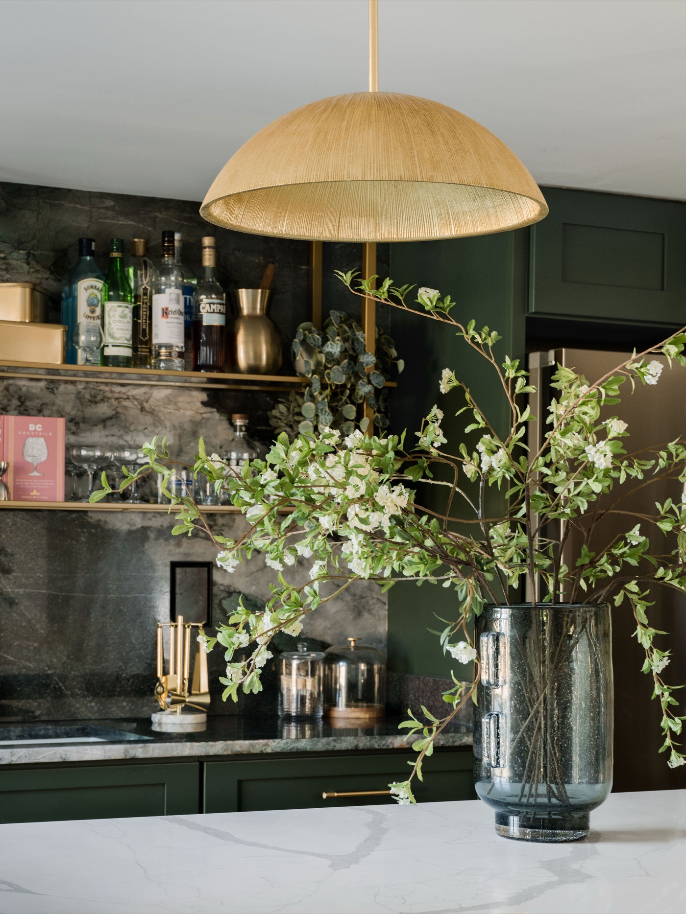 Brass bottle shelf and rail mounted on the antiqued back wall of the green wet bar beneath a woven dome pendant in the Takoma Park project by Jennifer Kizzee Design