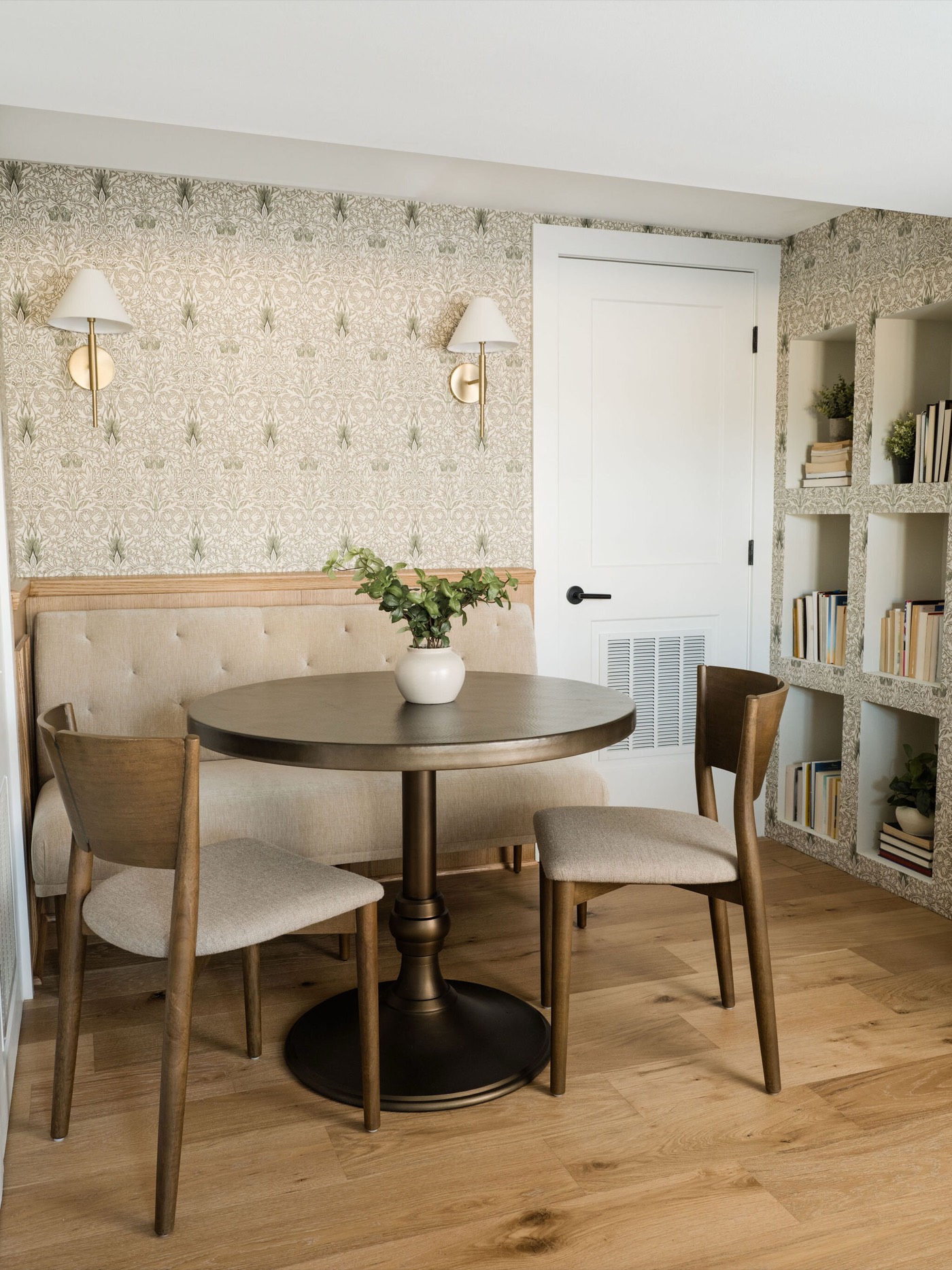 Breakfast nook with a cream tufted banquette, a round pedestal table, and two brass sconces on sprig patterned wallpaper, in the Takoma Park project by Jennifer Kizzee Design