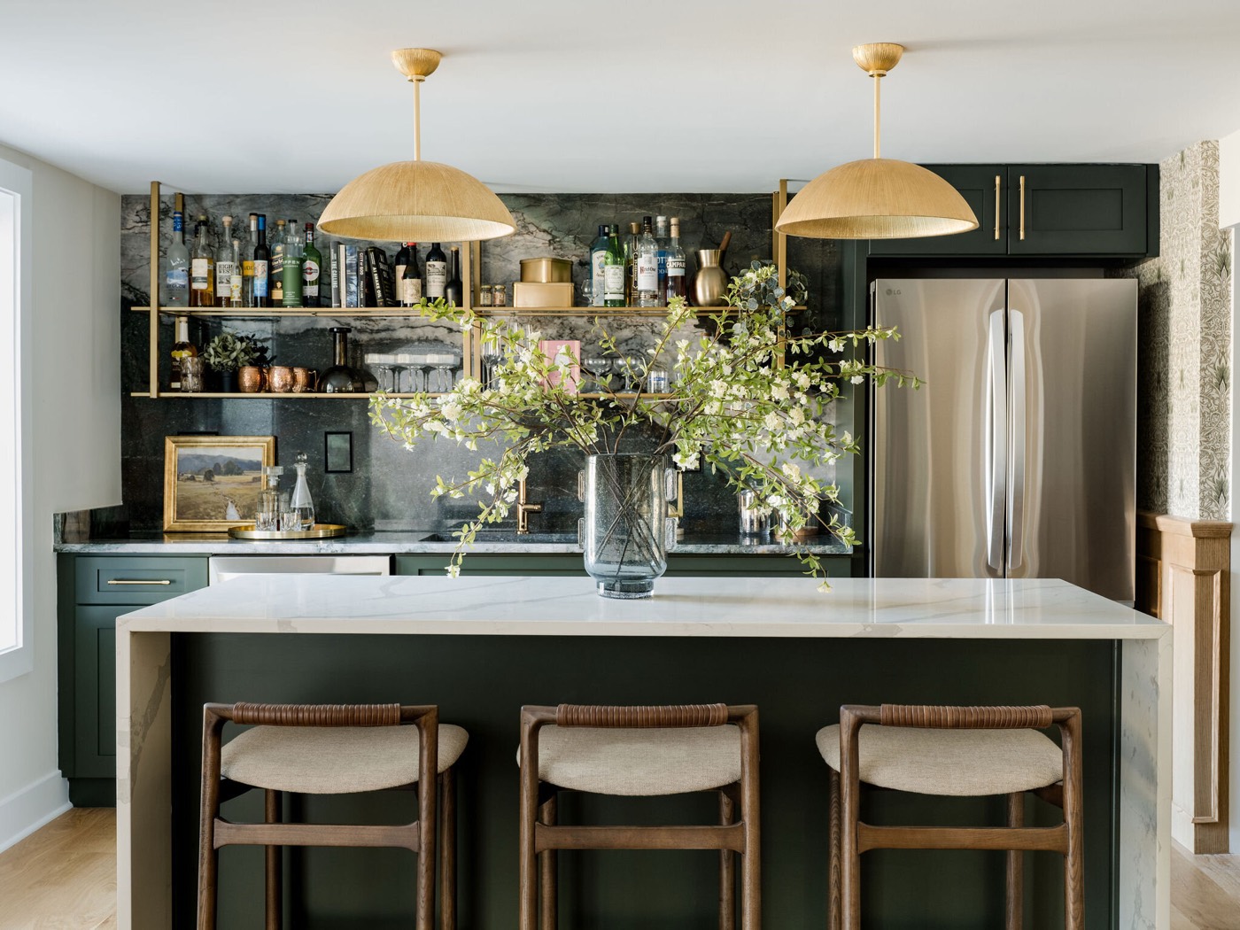 Wet bar in deep green cabinetry with a brass bottle shelf on an antiqued back wall, under two dome pendants above a white stone island, in the Takoma Park project by Jennifer Kizzee Design
