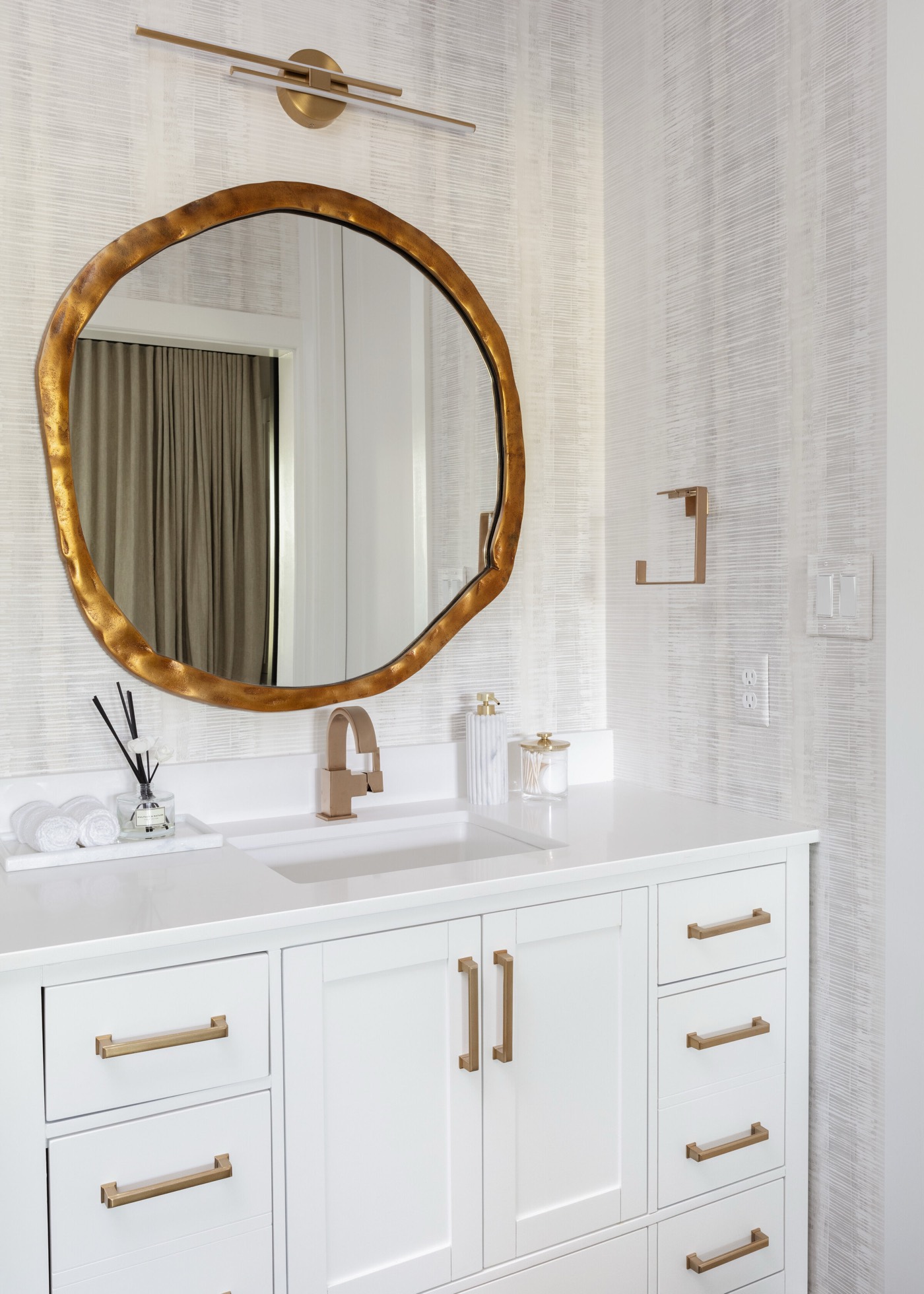 Bath vanity with an organic-edge gold mirror under a brass linear light and a brass faucet in the Vue Point project by Jennifer Kizzee Design