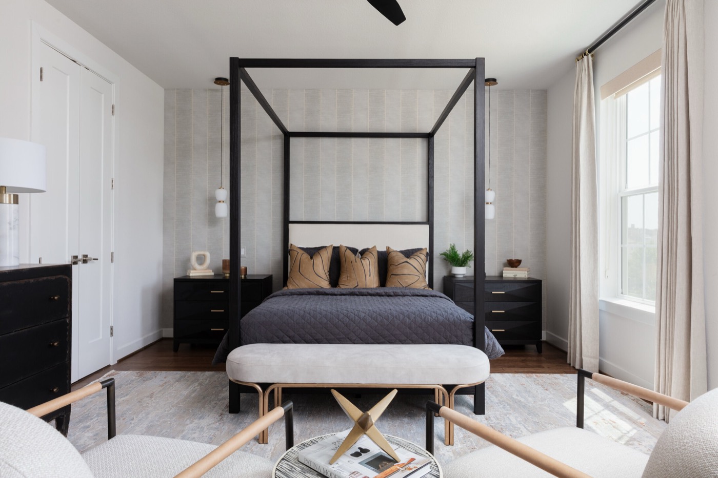 Bedroom with a black steel canopy bed, twin white glass bedside pendants, and a cream bench on brass legs in the Vue Point project by Jennifer Kizzee Design