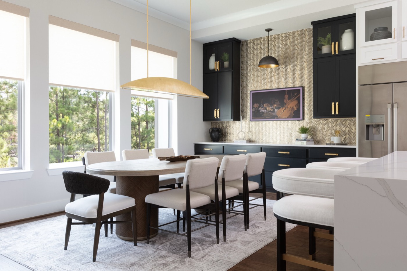 Dining room with an oval wood table under a brass linear pendant, set against a navy cabinet wall with a gold herringbone panel in the Vue Point project by Jennifer Kizzee Design